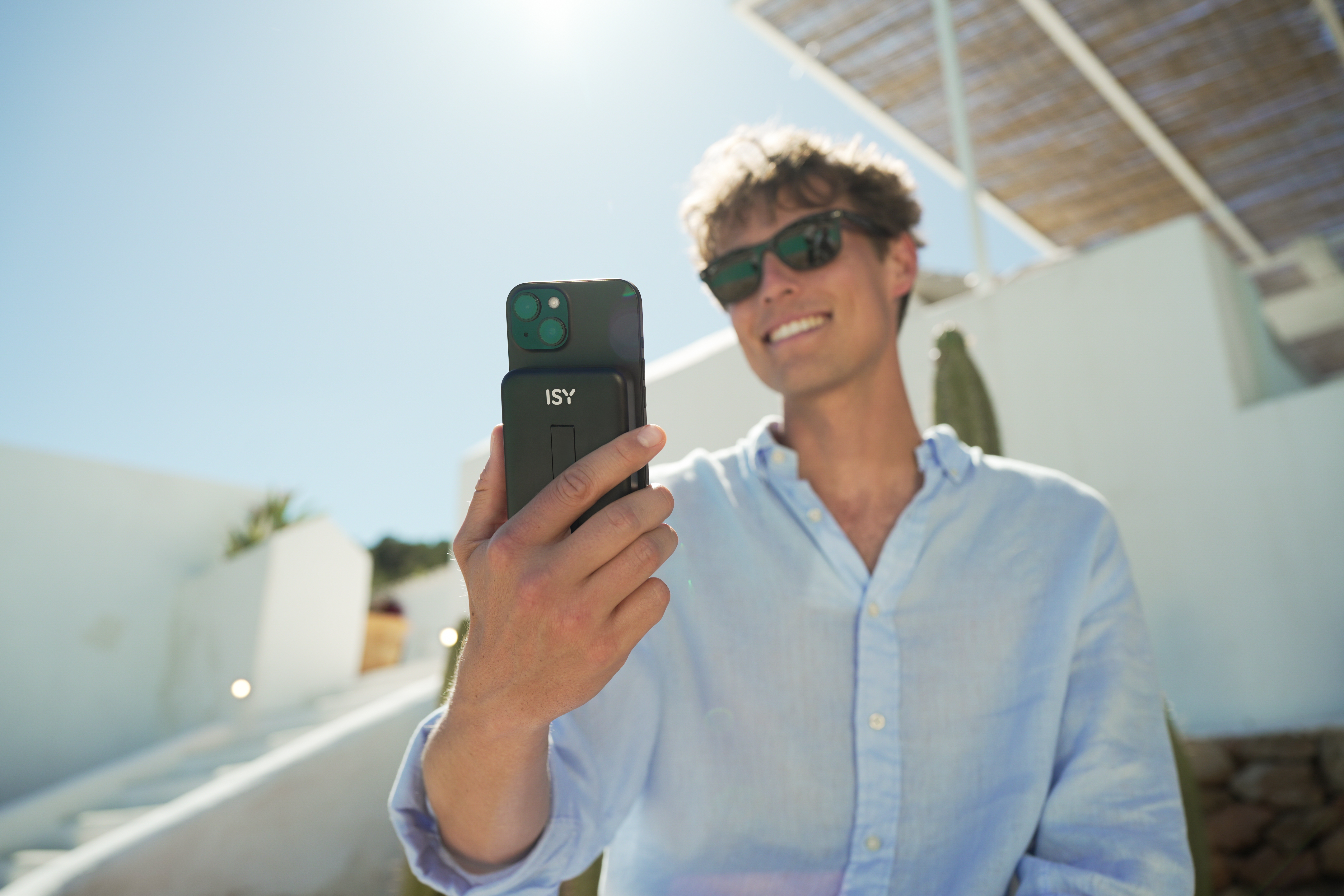 Ein Mann macht ein Selfie mit seinem Handy vor einem weißen Gebäude bei strahlendem Sonnenschein.