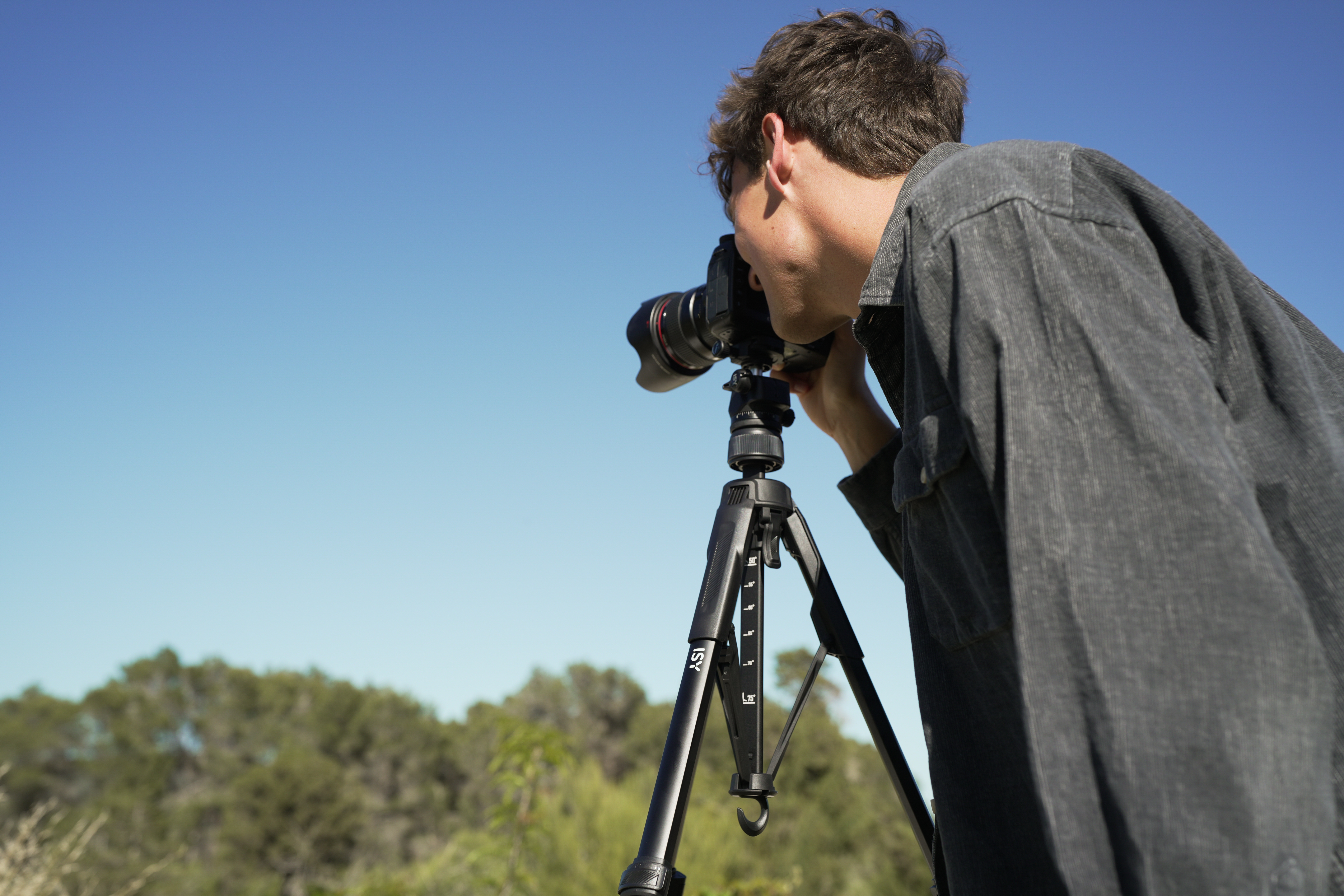 Ein Mann im grauen Hemd fotografiert mit einer Kamera auf einem Stativ unter klarem Himmel.