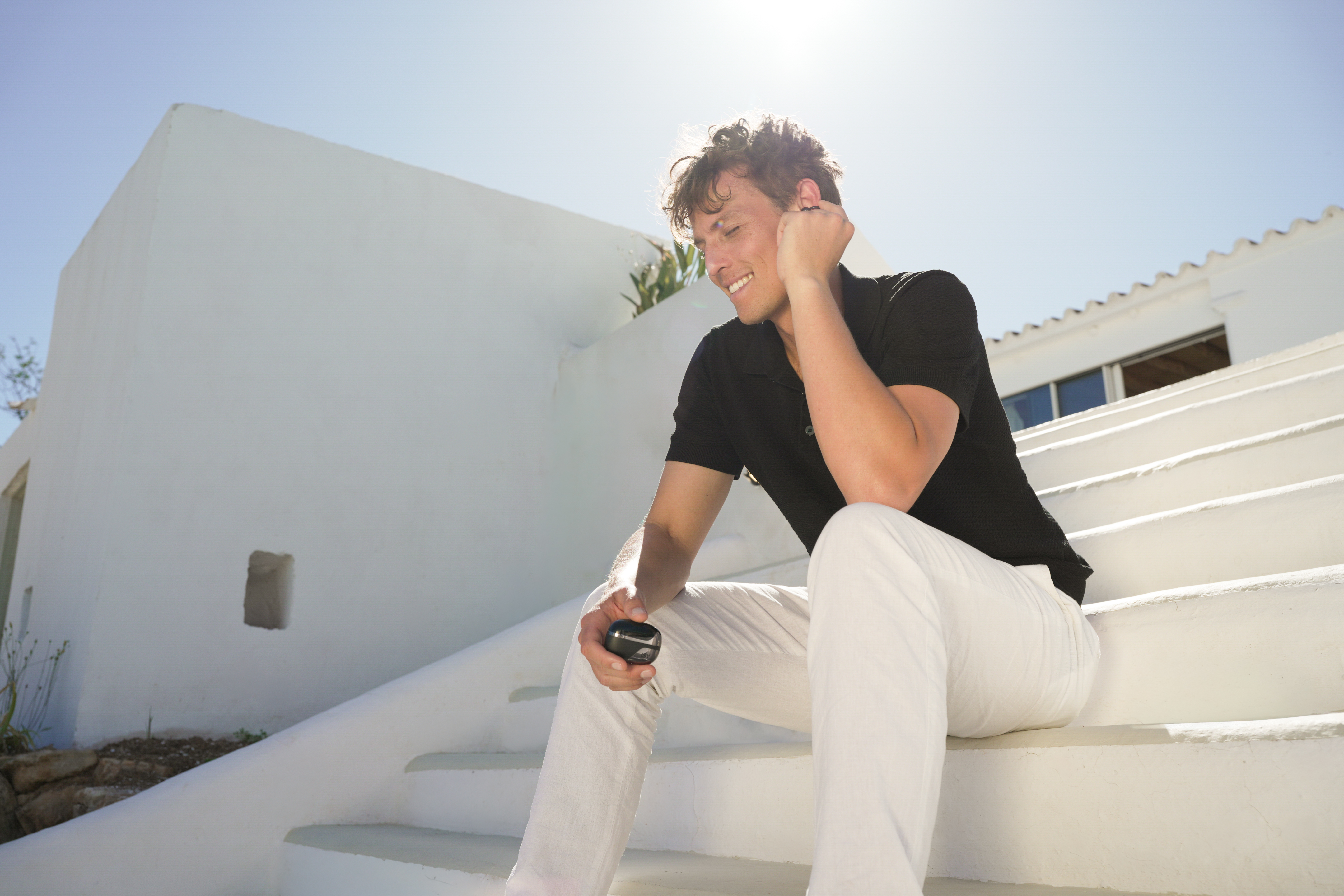 Mann in schwarzem Poloshirt und weißer Hose sitzt auf weißen Stufen mit blauem Himmel.