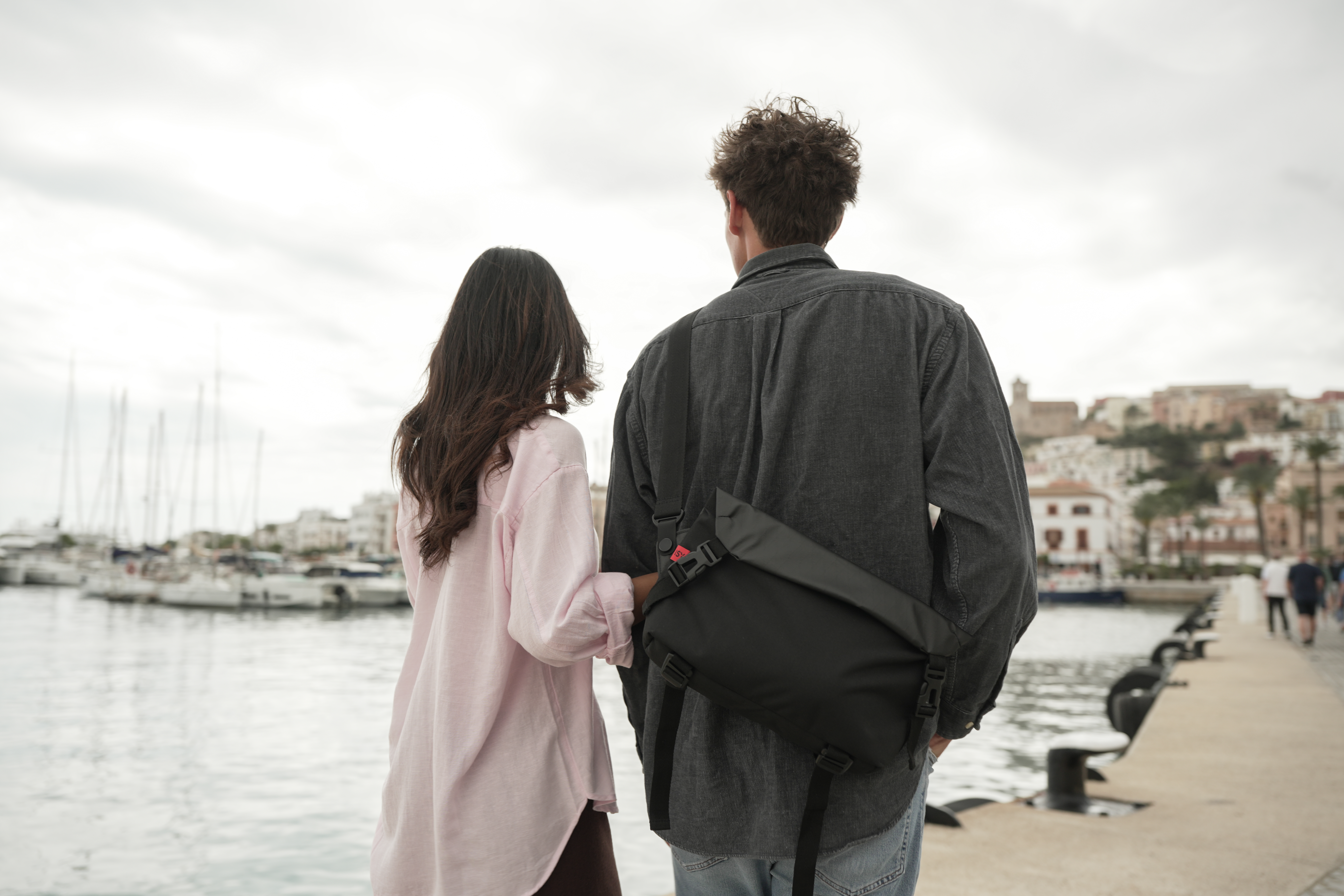 Ein Mann und eine Frau stehen am Kai mit Blick auf Boote und eine Stadt.