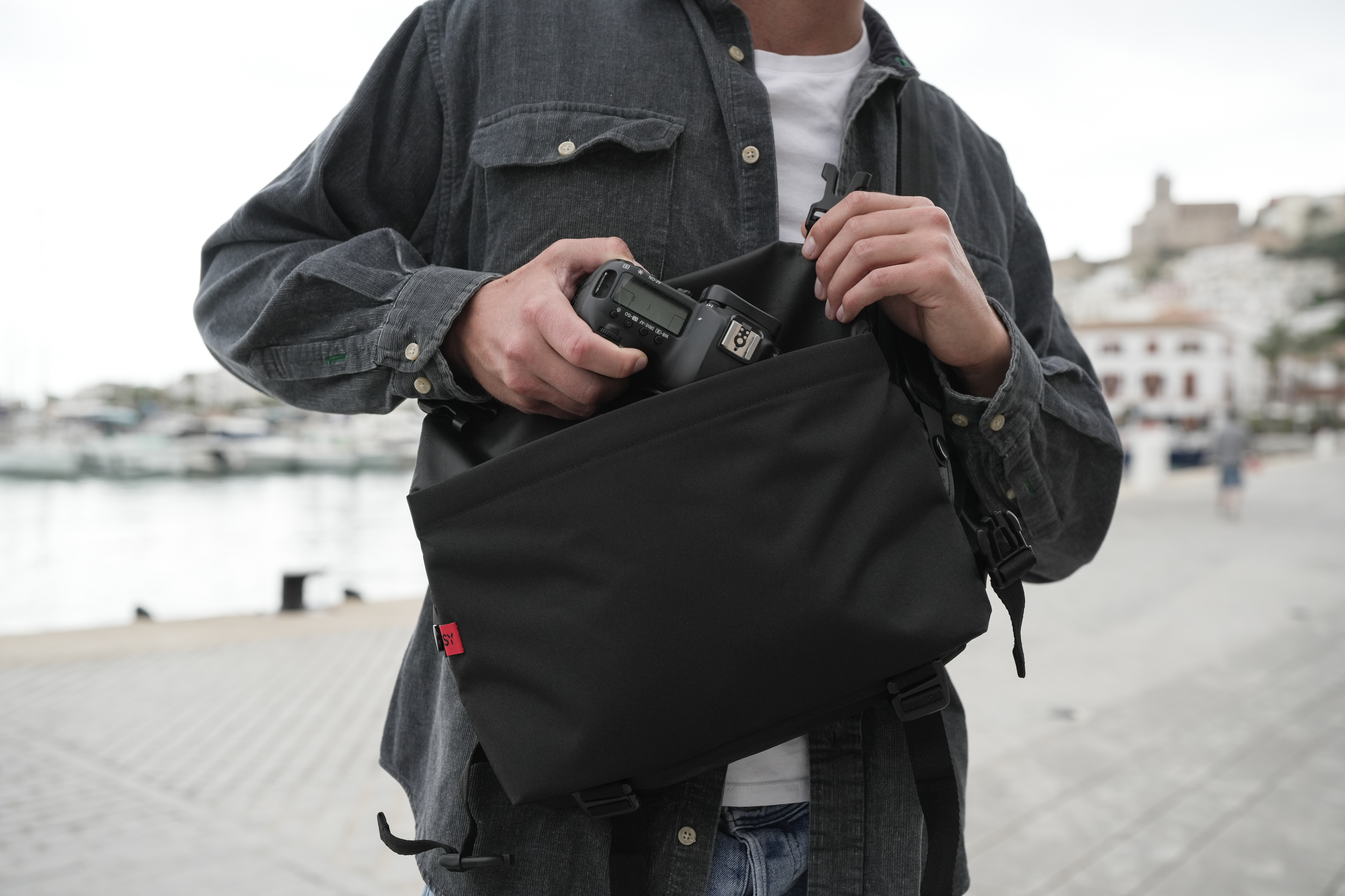 Mann mit dunkler Jacke packt Kamera in schwarze Tasche, Hafen im Hintergrund.