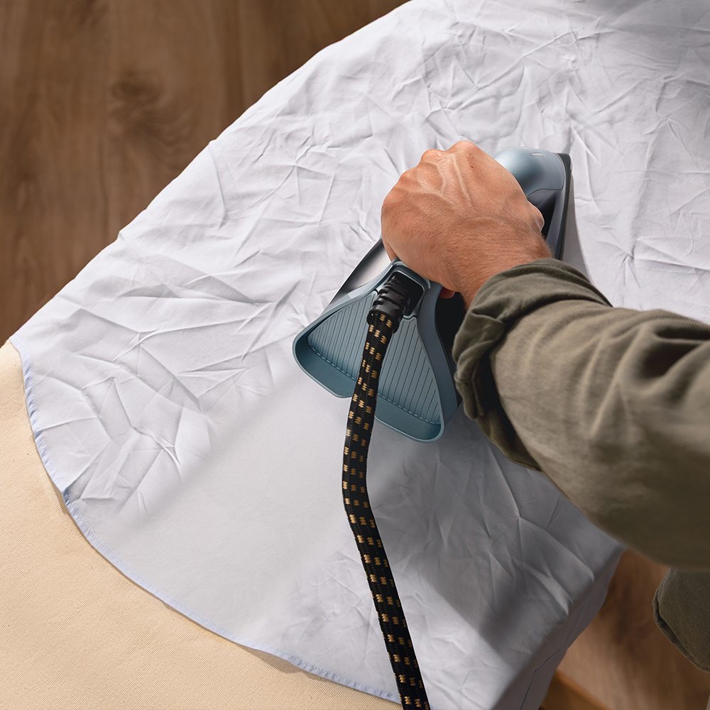 Una mano sostiene una plancha sobre una camisa blanca arrugada en una tabla de planchar.