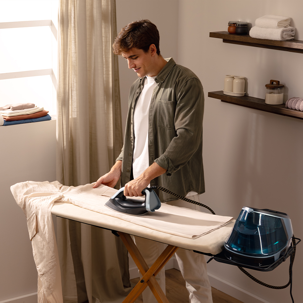 Hombre plancha pantalones en tabla de planchar. Plancha de vapor y depósito de agua cerca.