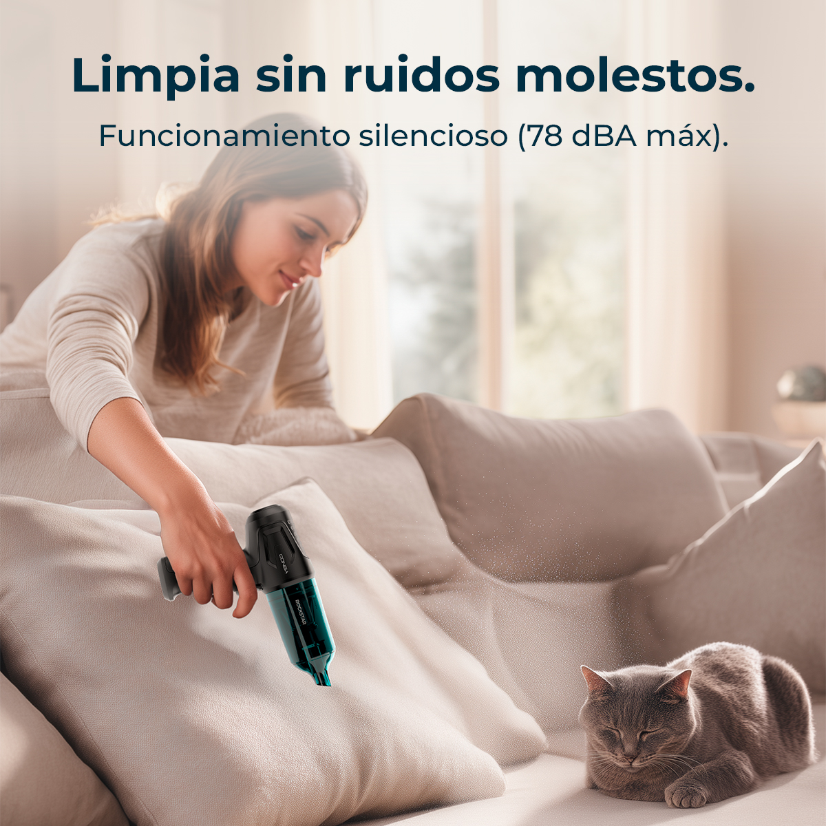 Mujer limpiando sofá con aspirador de mano, gato duerme cerca.