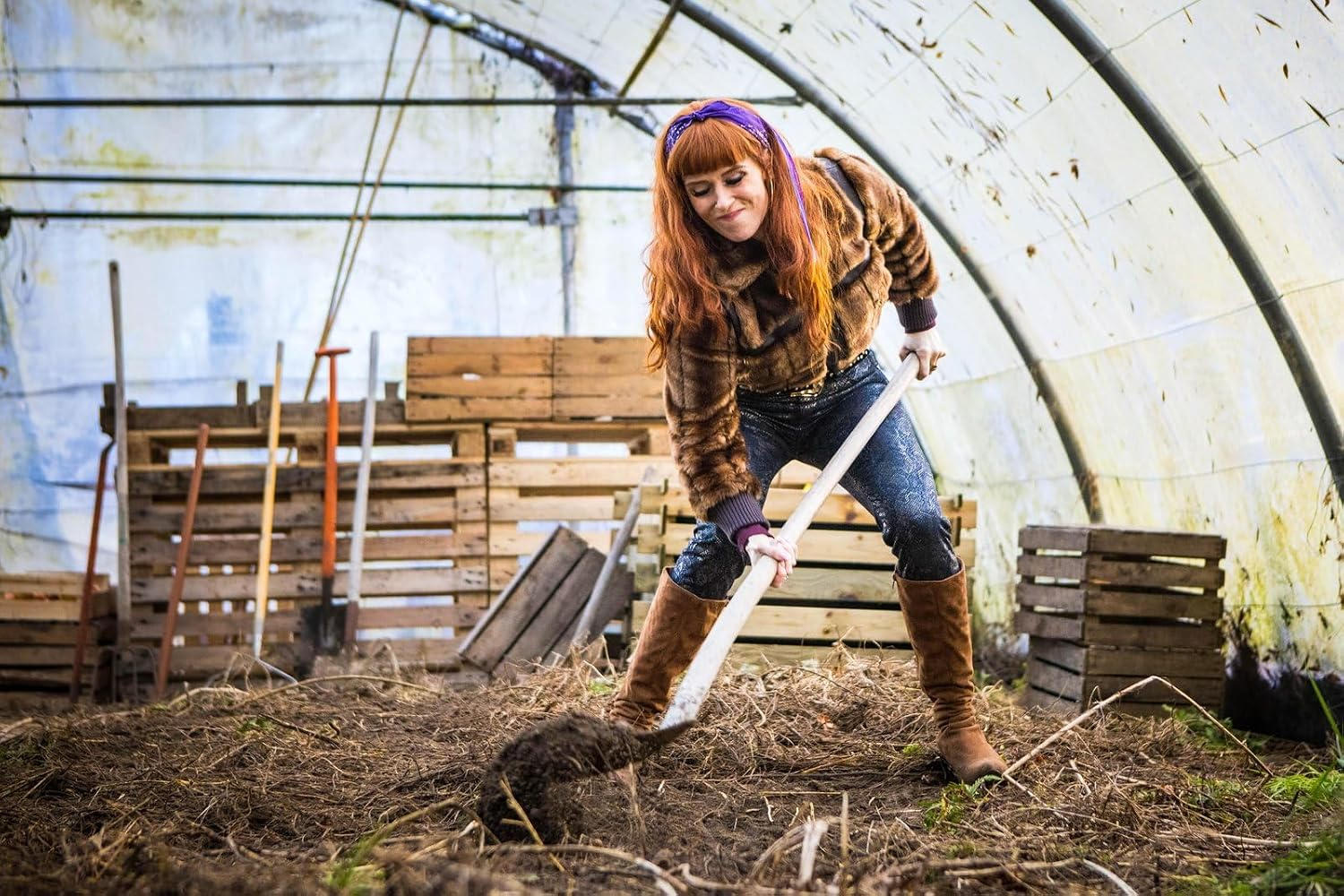 Frau mit roten Haaren gräbt in einem Gewächshaus mit Werkzeugen und Holzkisten.