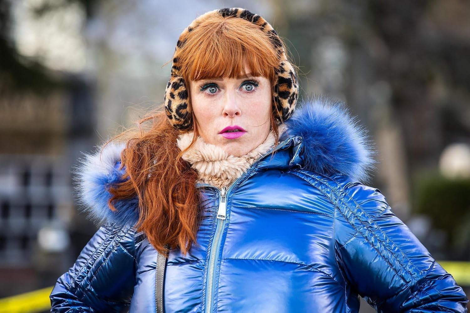Frau mit roten Haaren, blauer Daunenjacke, Leoparden-Ohrenschützern und pinkem Lippenstift.