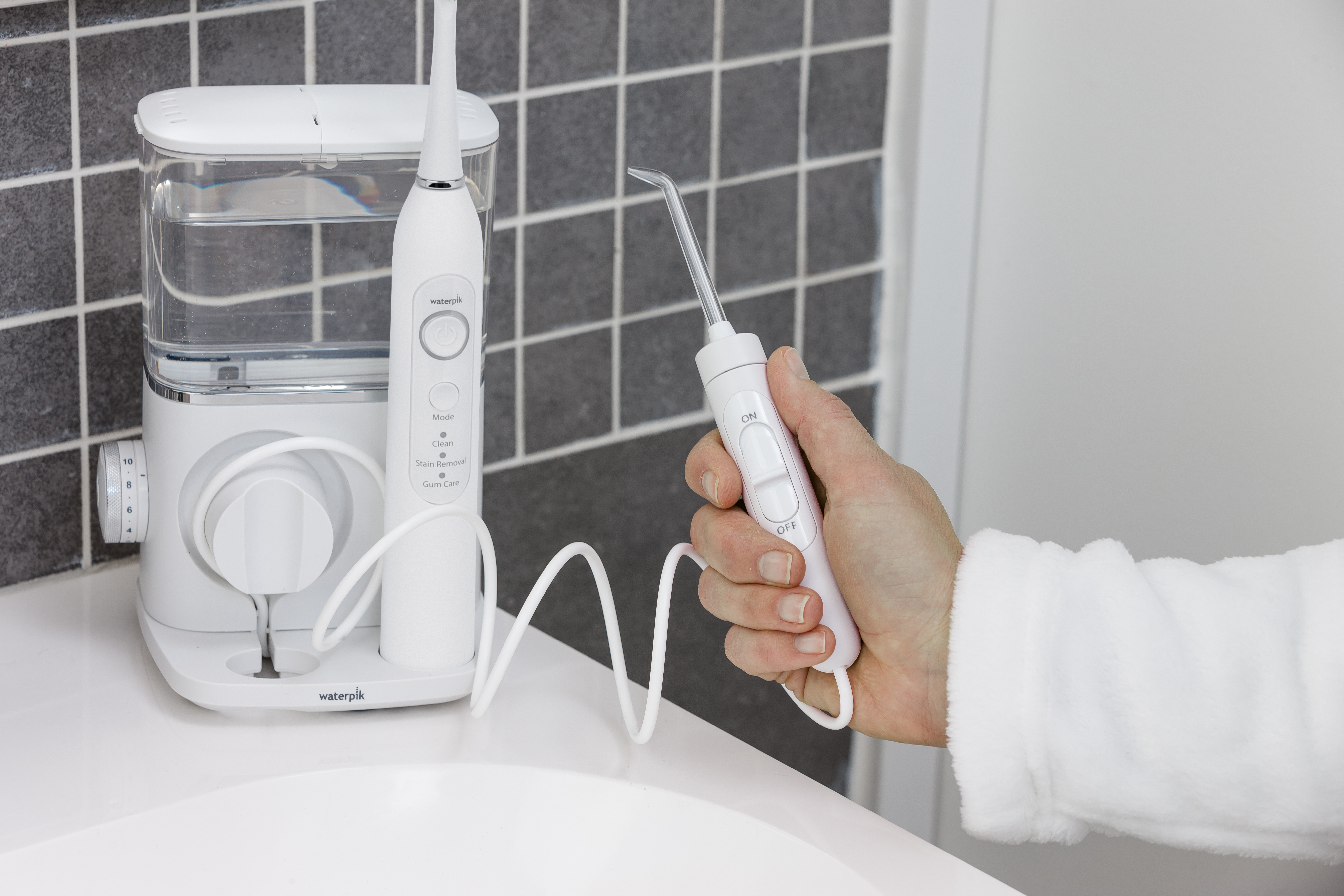 Biały uchwyt irygatora Waterpik trzyma dłoń w białym szlafroku.
