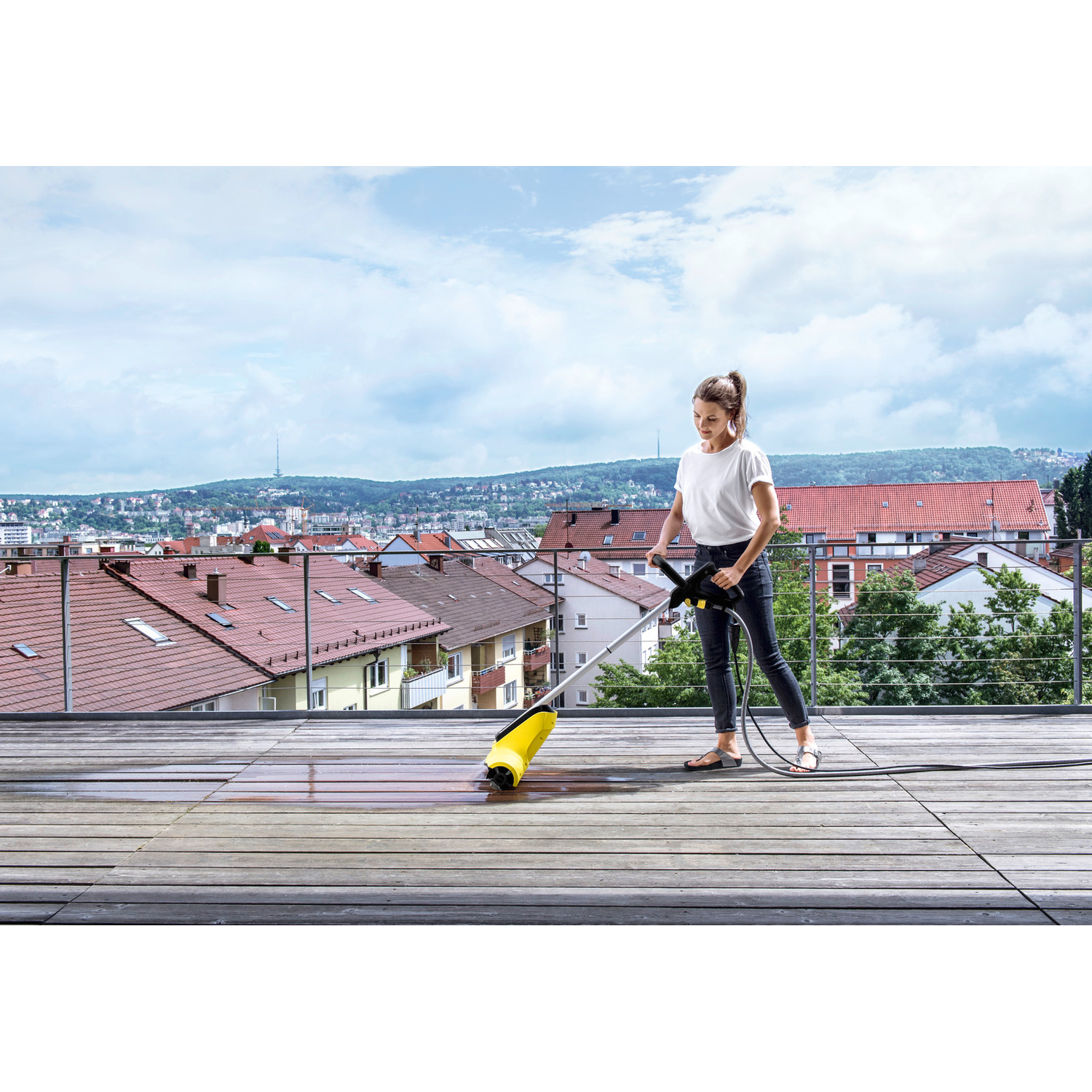 Donna pulisce terrazza in legno con idropulitrice, città sullo sfondo.