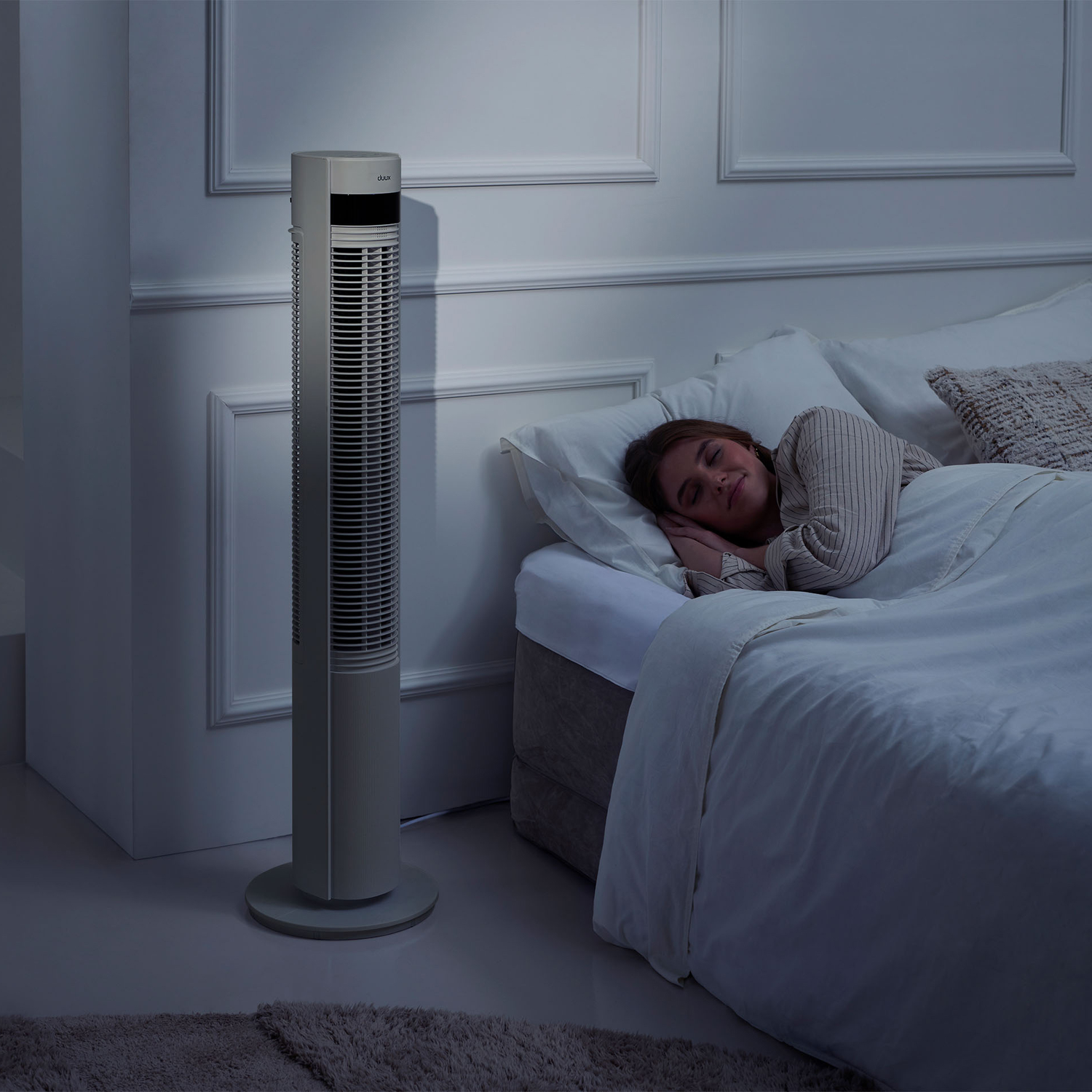 Een slanke torenventilator staat naast een bed waar een vrouw slaapt.