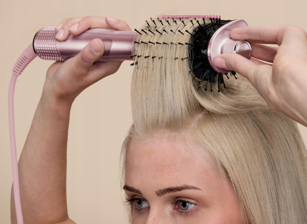 Osoba stylizuje blond włosy za pomocą różowej lokówki i okrągłej szczotki.