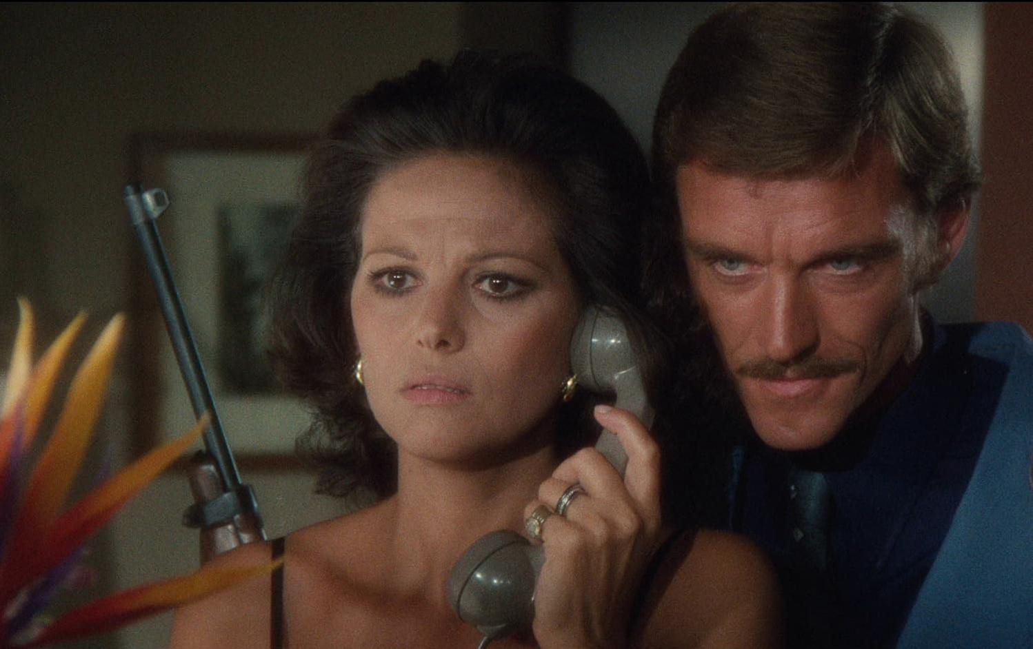 Eine Frau mit einem Telefonhörer und ein Mann mit Schnurrbart, im Hintergrund eine Blume und ein Gewehr.
