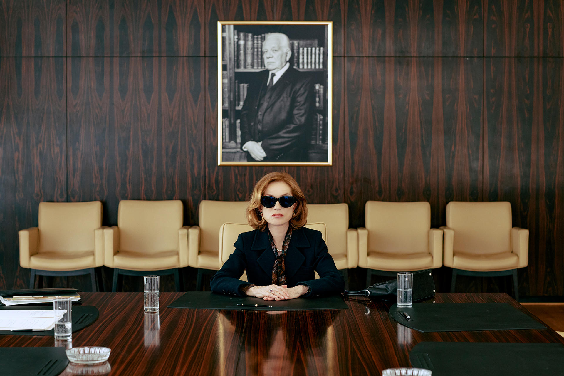Frau mit Sonnenbrille sitzt am Konferenztisch unter einem Porträt eines Mannes.