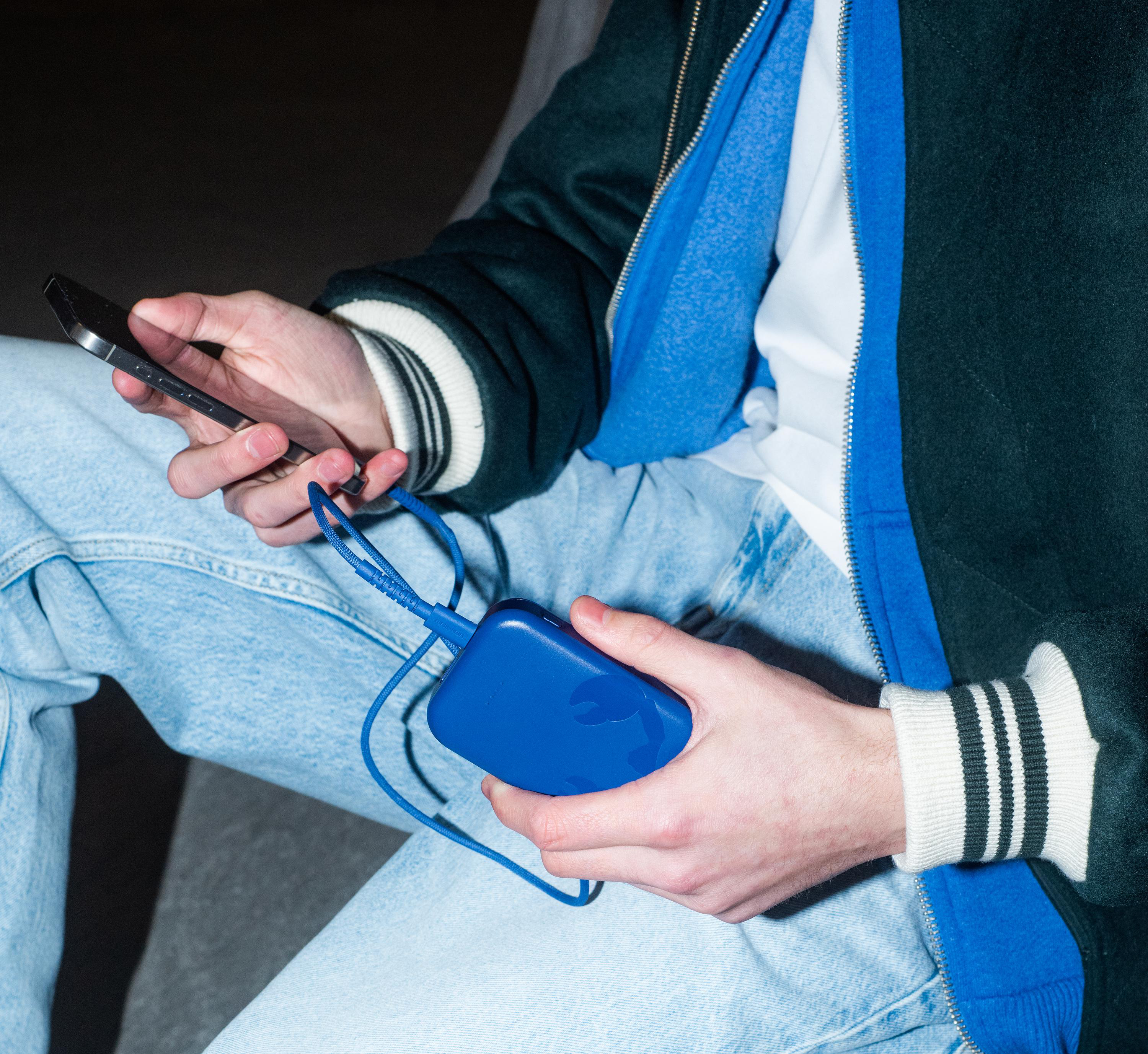 Person lädt Handy mit Powerbank auf, trägt Jeans und dunkle Jacke.