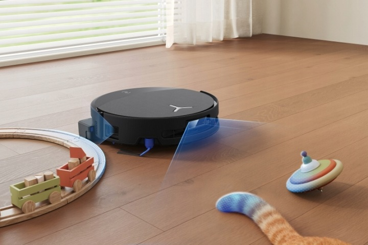 Czarny robot sprzątający porusza się po drewnianej podłodze wokół zabawek i ogona kota.