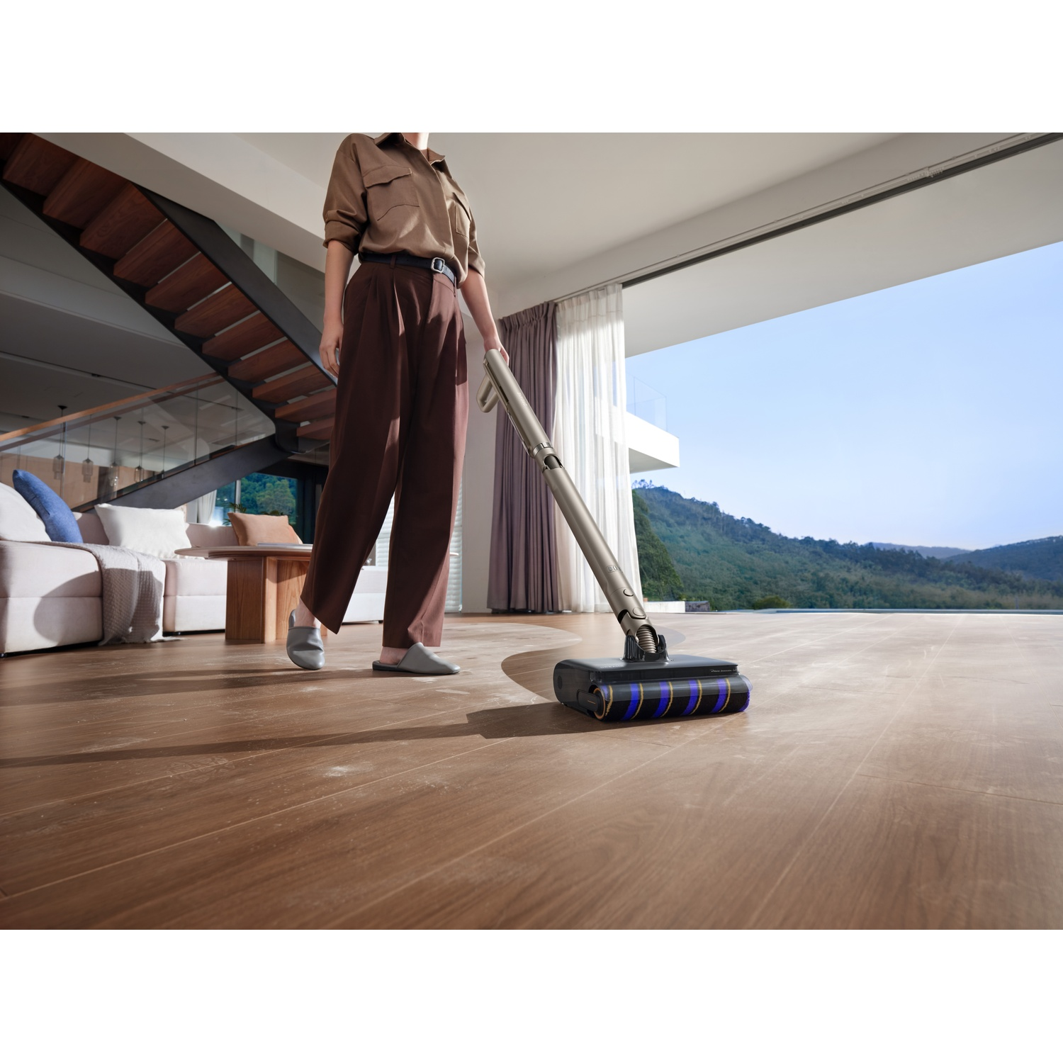 Persoon stofzuigt houten vloer met draadloze stofzuiger in moderne woonkamer met trap en uitzicht.