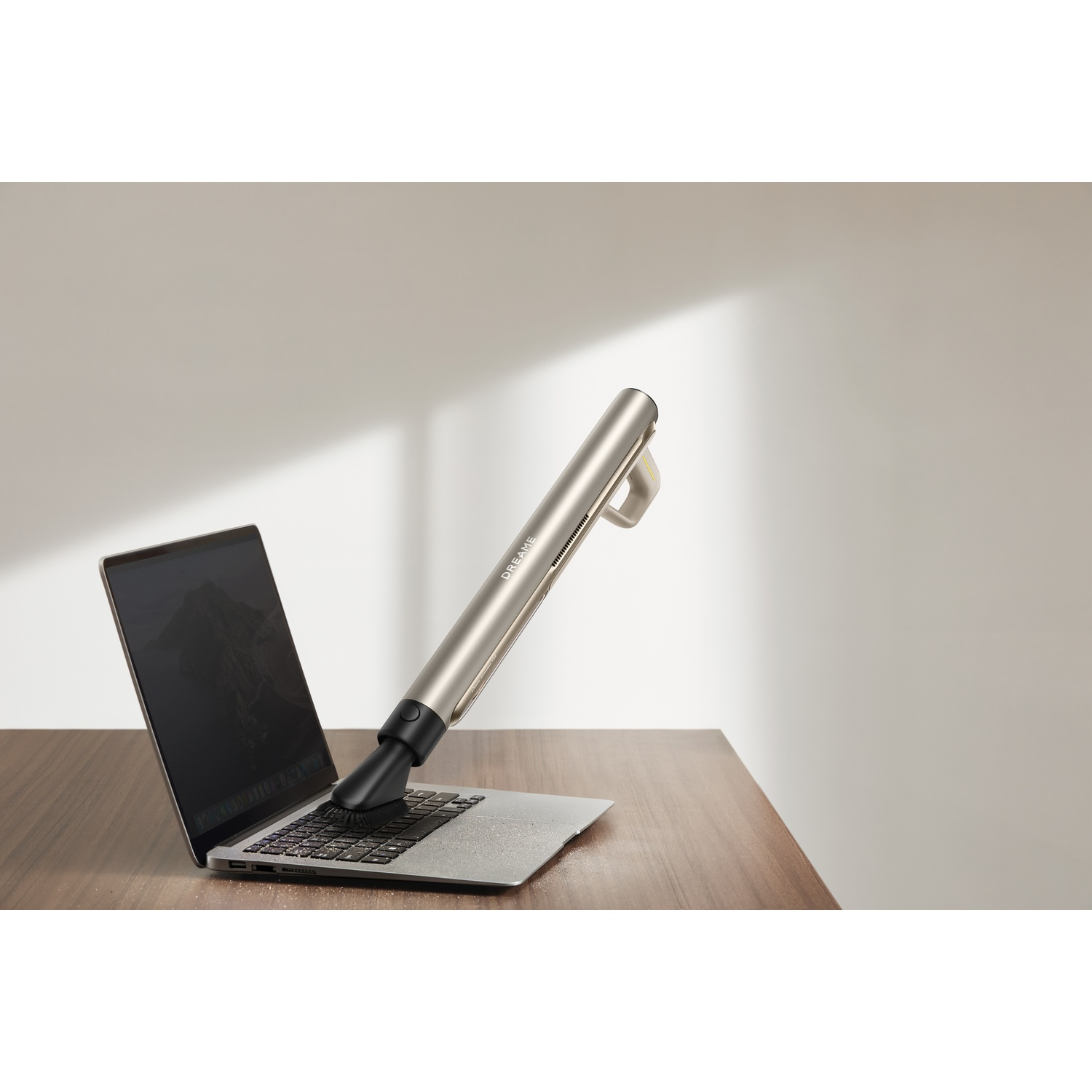 Een zilveren handstofzuiger reinigt het toetsenbord van een open laptop op een houten tafel.