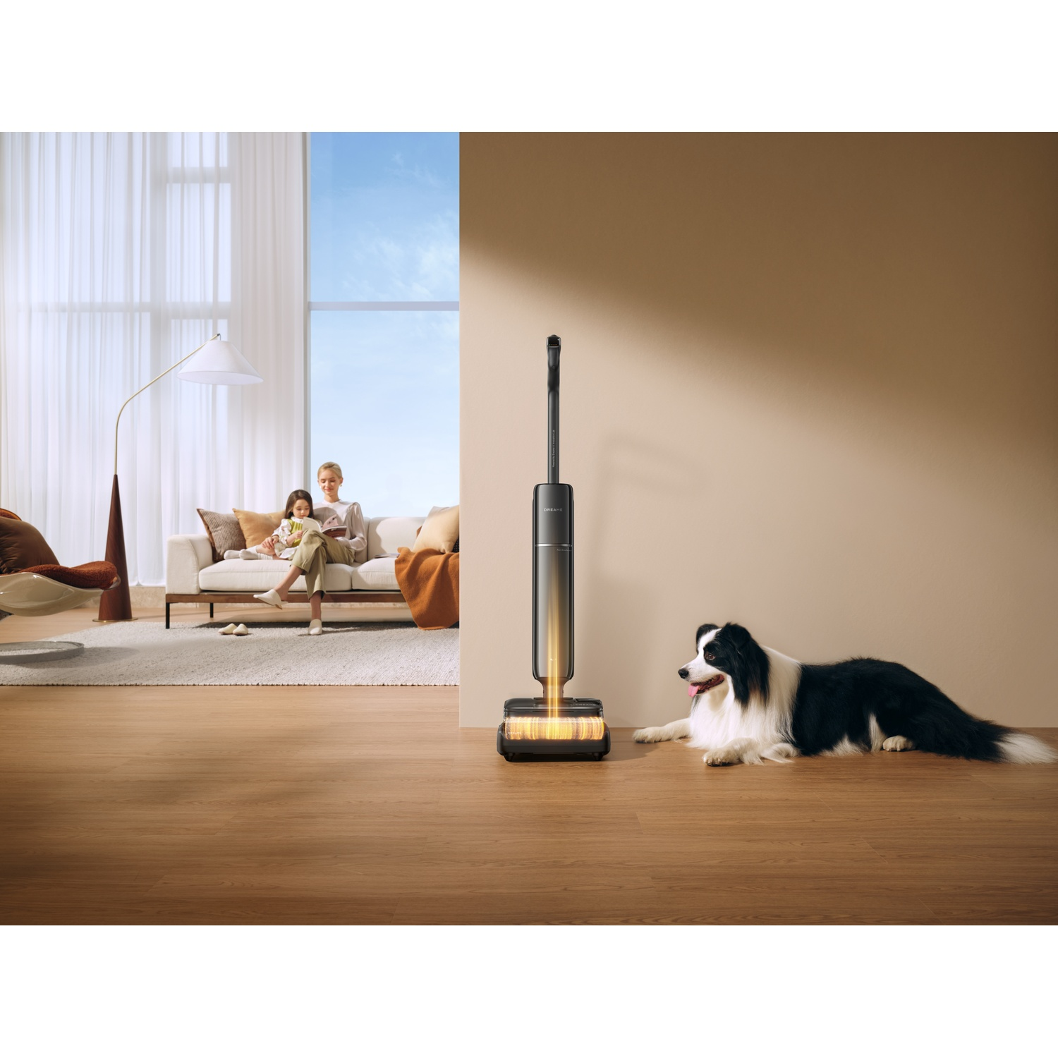 A black and white dog lies on a wooden floor next to a vacuum cleaner. A woman and child sit on a sofa.