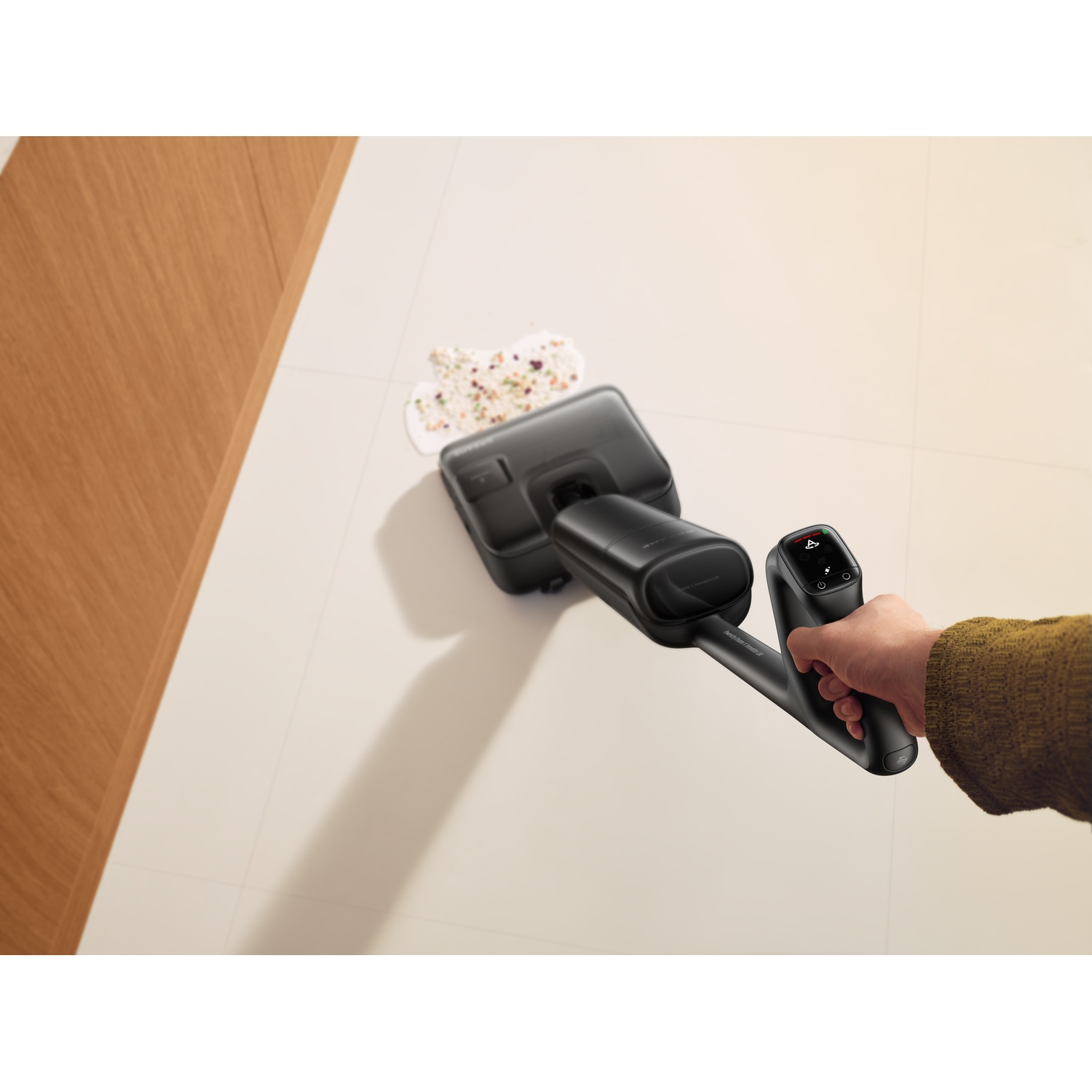 A hand holds a black vacuum cleaner, sucking up debris on a light-colored floor.