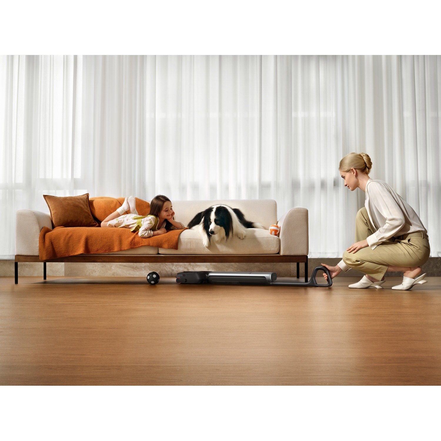 A woman kneels operating a vacuum cleaner while a girl and dog relax on a couch.