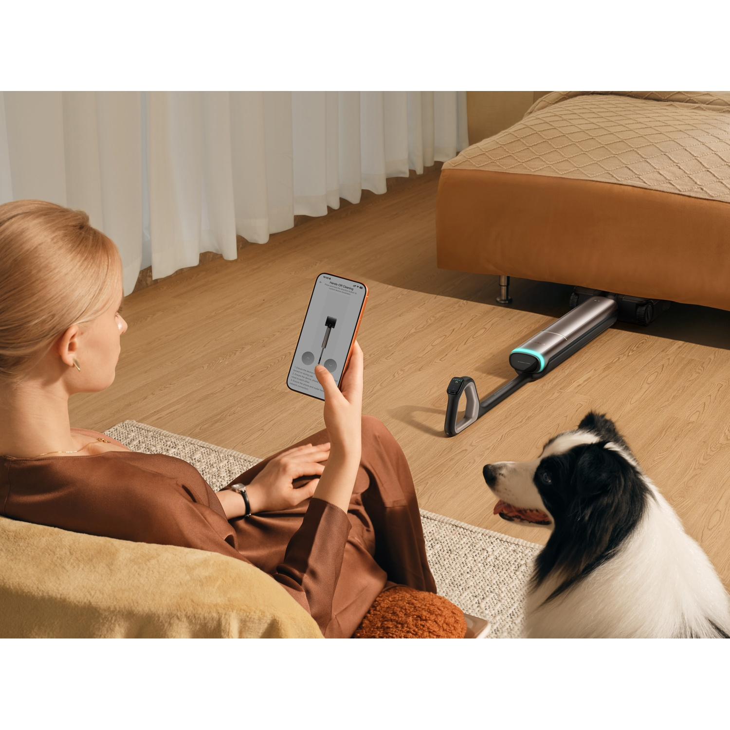 A woman sits on a sofa holding a smartphone. A robot vacuum is under a bed. A dog is nearby.