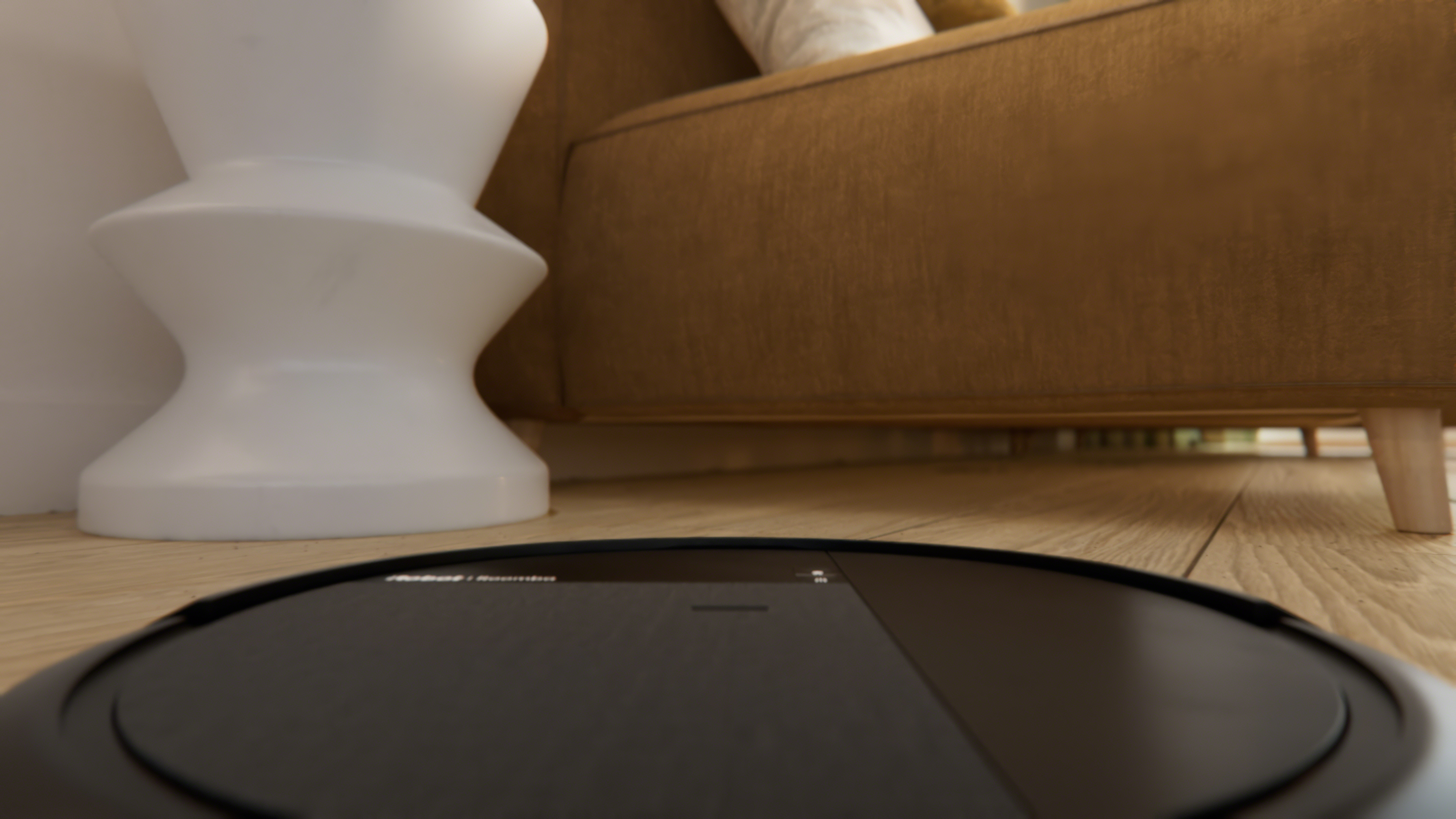Ein schwarzer Saugroboter auf einem Holzboden vor einem braunen Sofa.