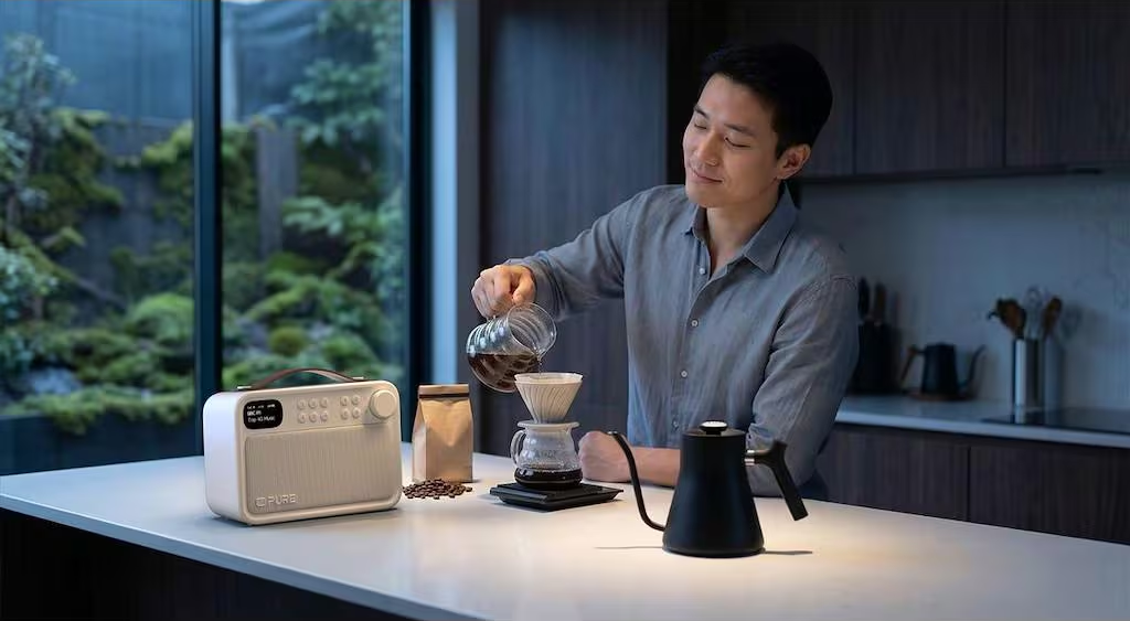 Un homme verse du café dans un filtre sur une balance, avec radio et bouilloire à proximité.