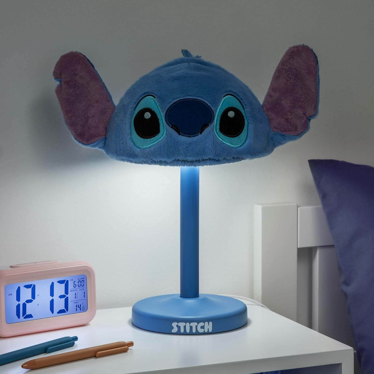 Kék Stitch lámpa világít meg egy éjjeliszekrényt rózsaszín ébresztőórával és tollakkal.
