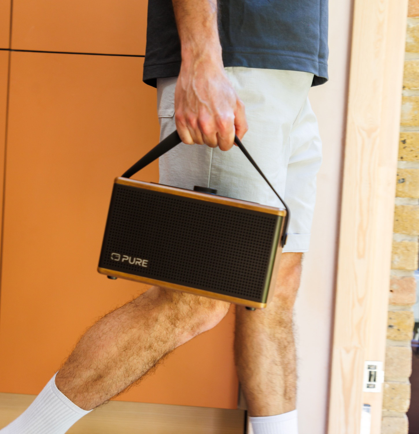 Mann mit dunklem T-Shirt und Shorts trägt eine schwarze Lautsprecherbox mit Holzrahmen.