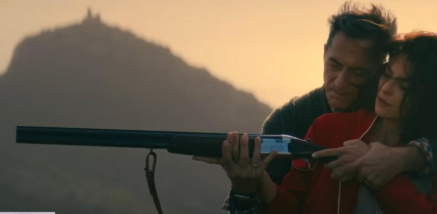 Mann und Frau mit Gewehr vor bergiger Landschaft im Sonnenuntergang.
