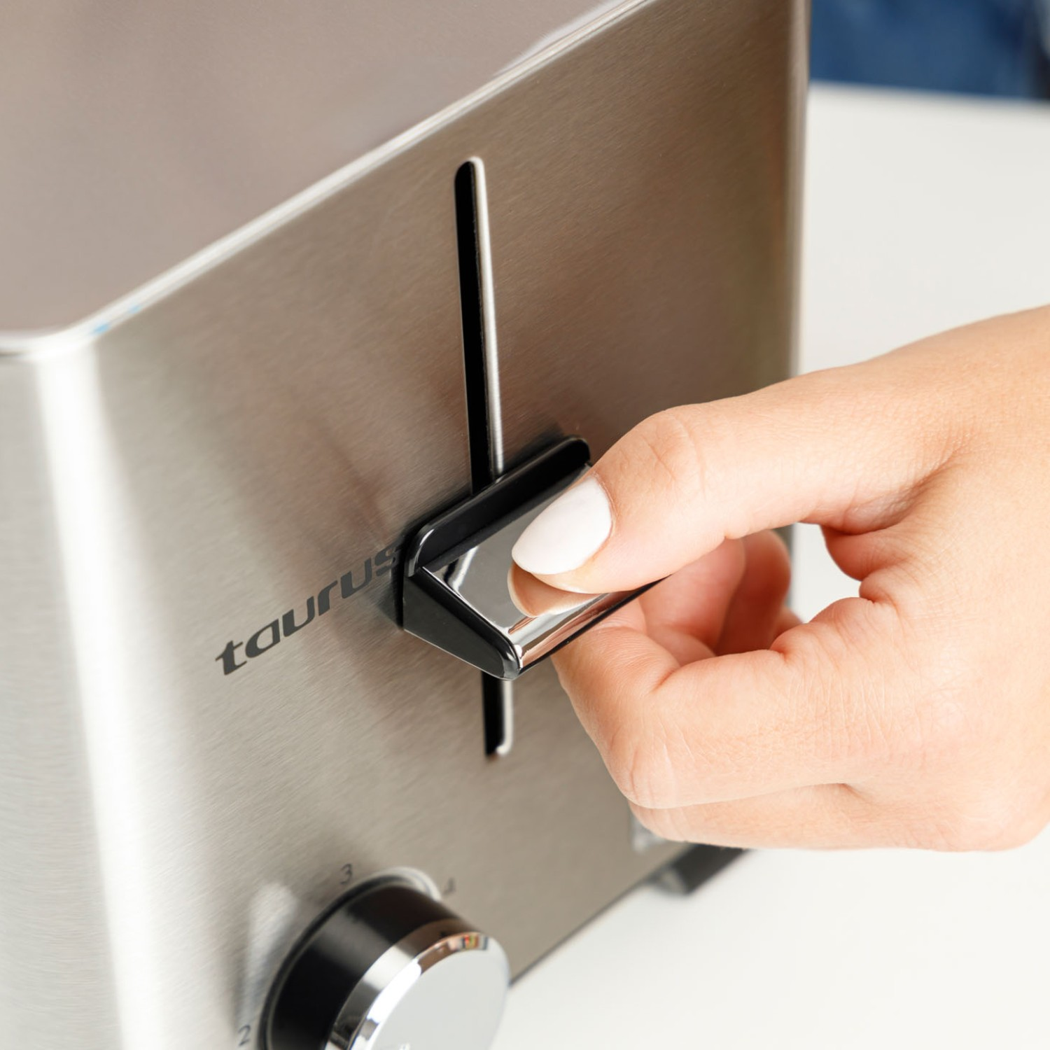 Una mano con uñas blancas presiona la palanca de una tostadora Taurus plateada.