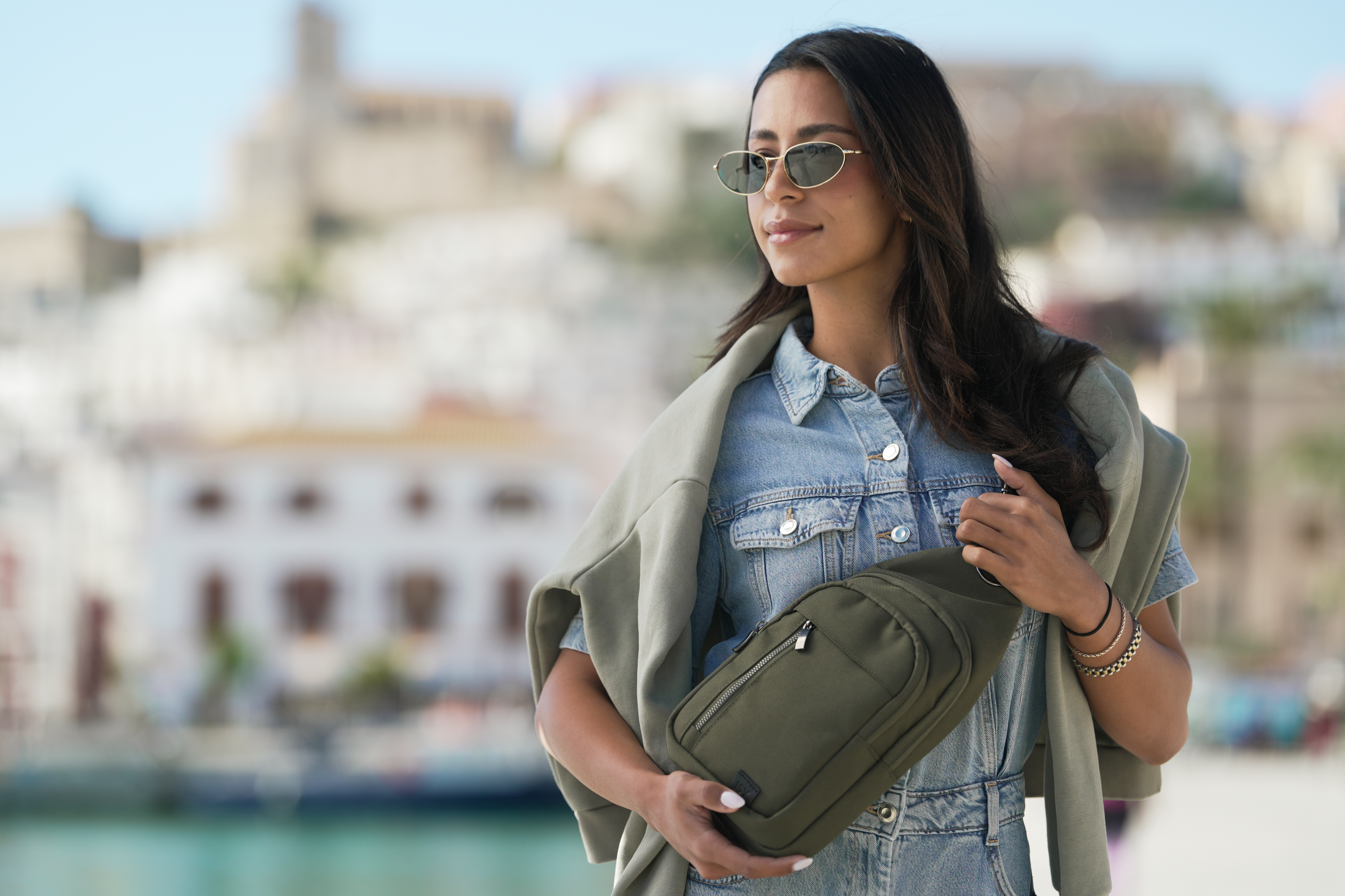 Eine Frau mit Sonnenbrille und Jeansjacke trägt eine grüne Umhängetasche. Im Hintergrund eine Stadt.