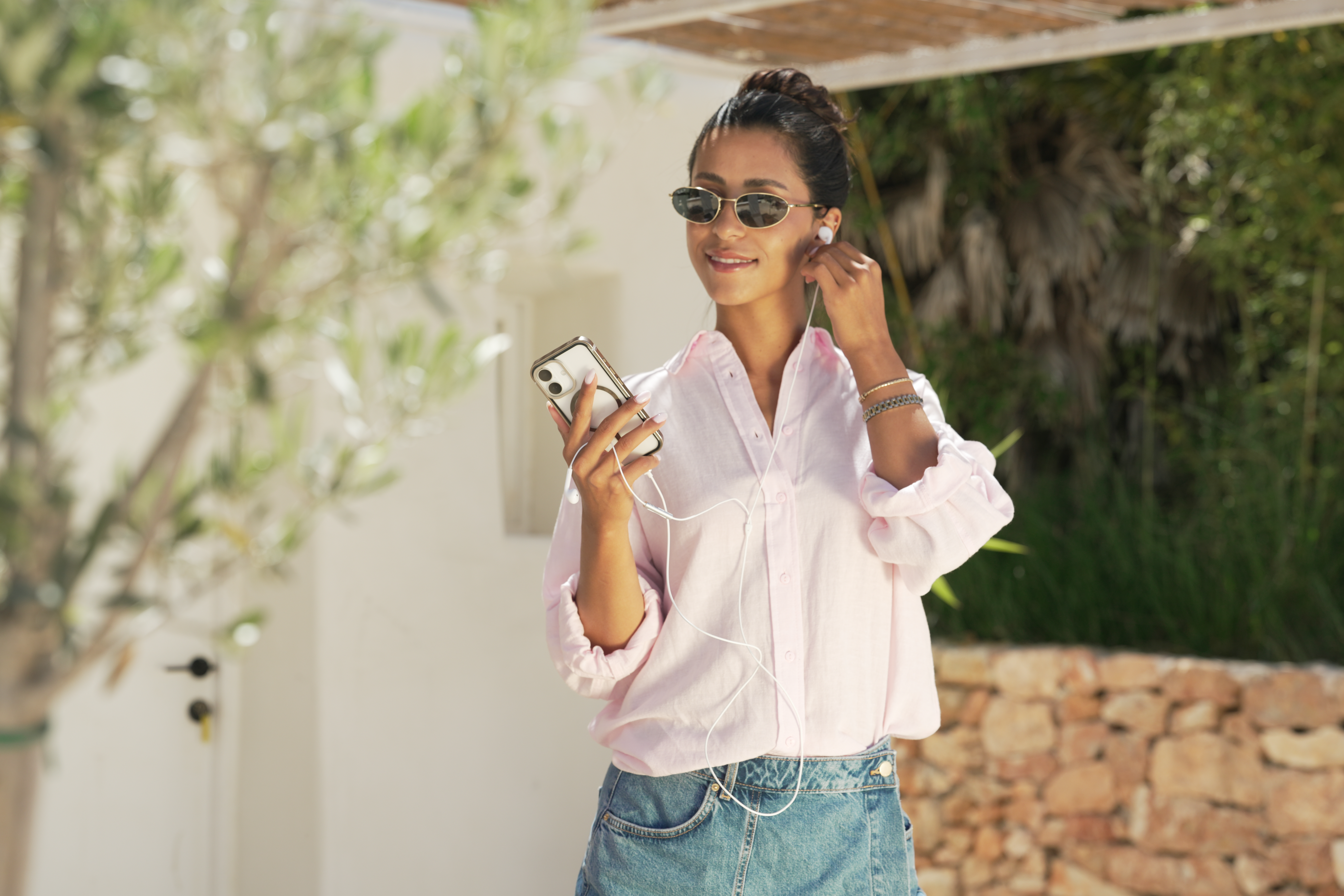 Frau im rosa Hemd, Jeansrock, Sonnenbrille, hört draußen Musik auf dem Handy.