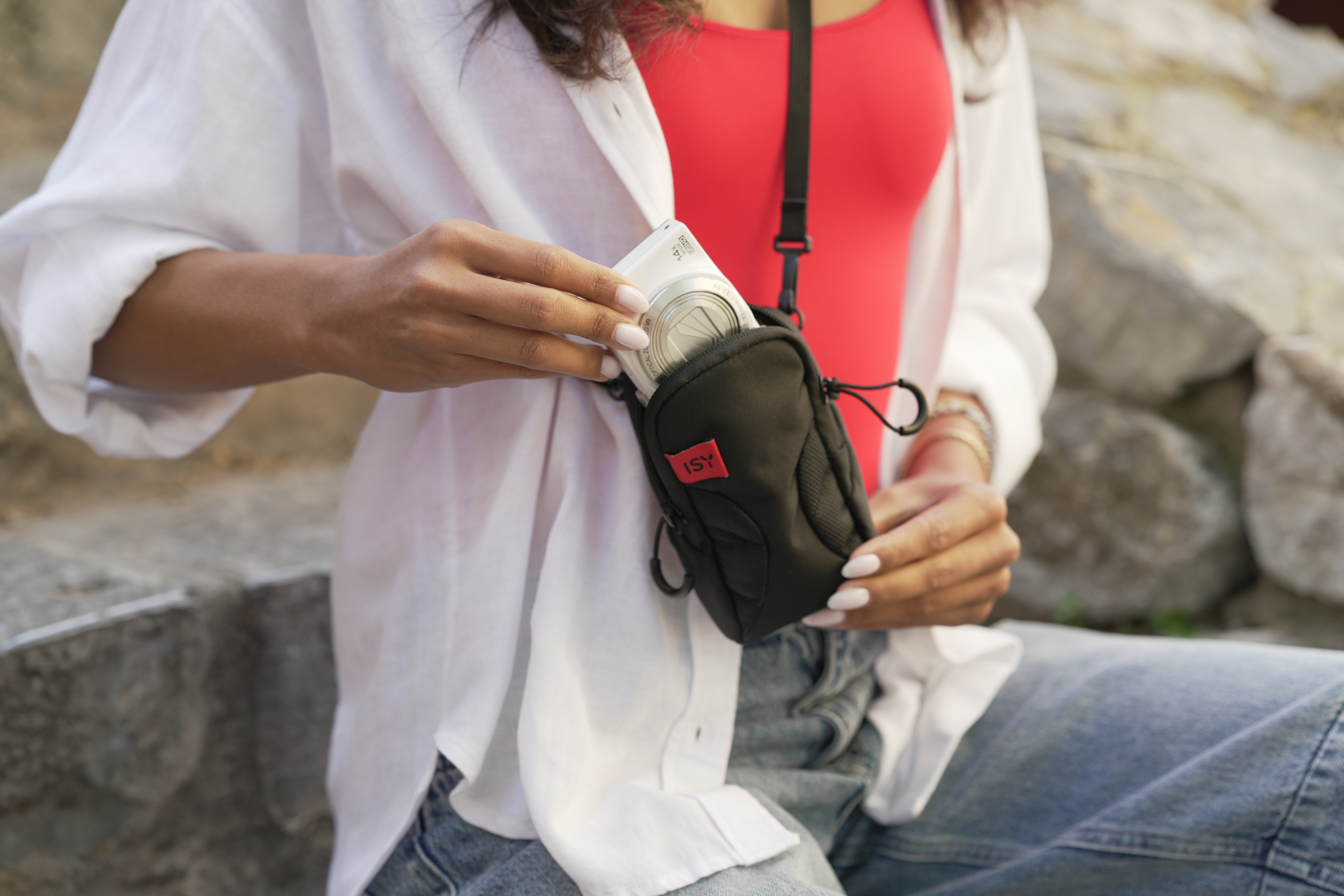 Eine Person legt eine weiße Kamera in eine schwarze Tasche. Sie trägt ein weißes Hemd und Jeans.