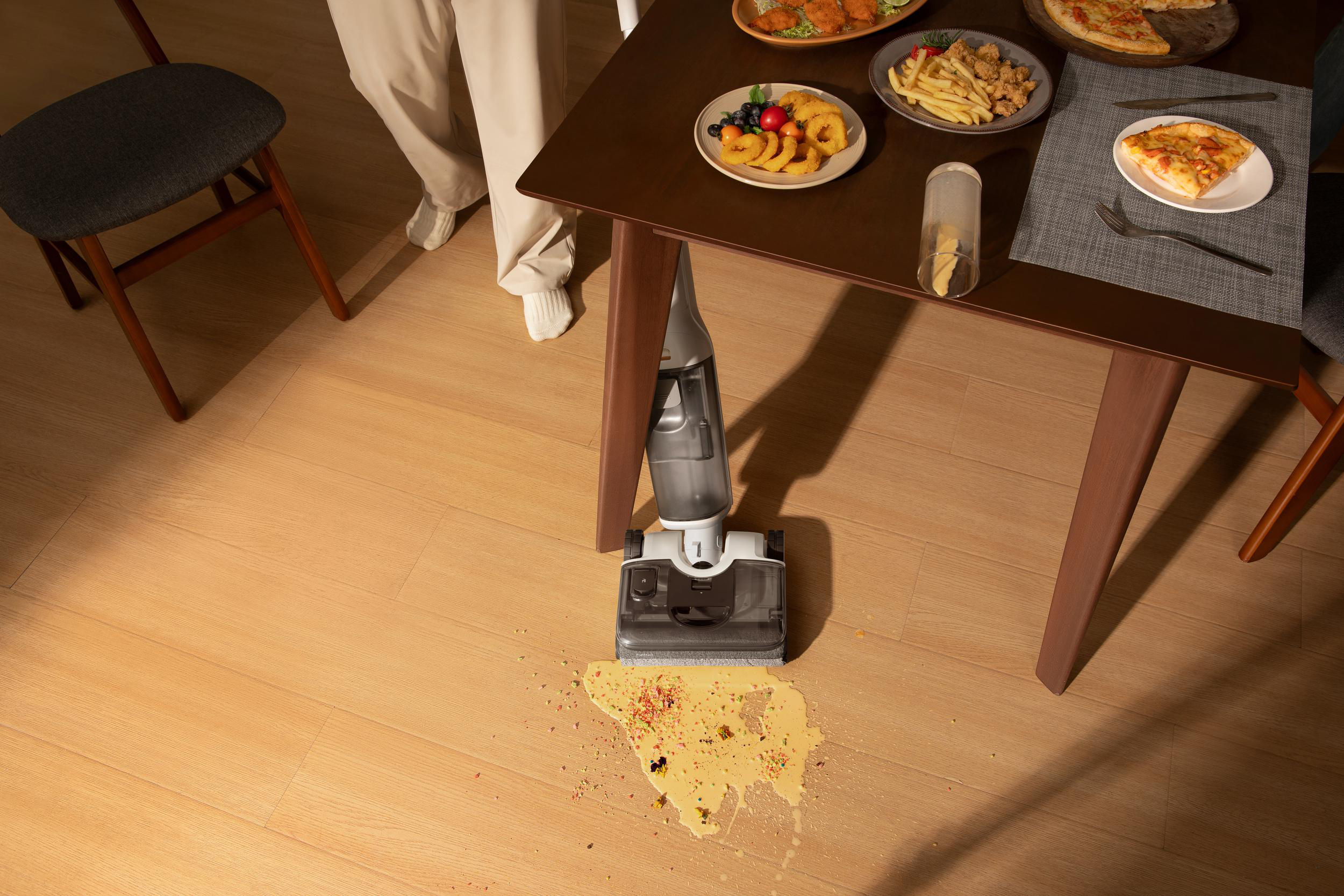 Ein Nass-/Trockensauger reinigt verschüttetes Essen auf einem Holzboden neben einem Esstisch.