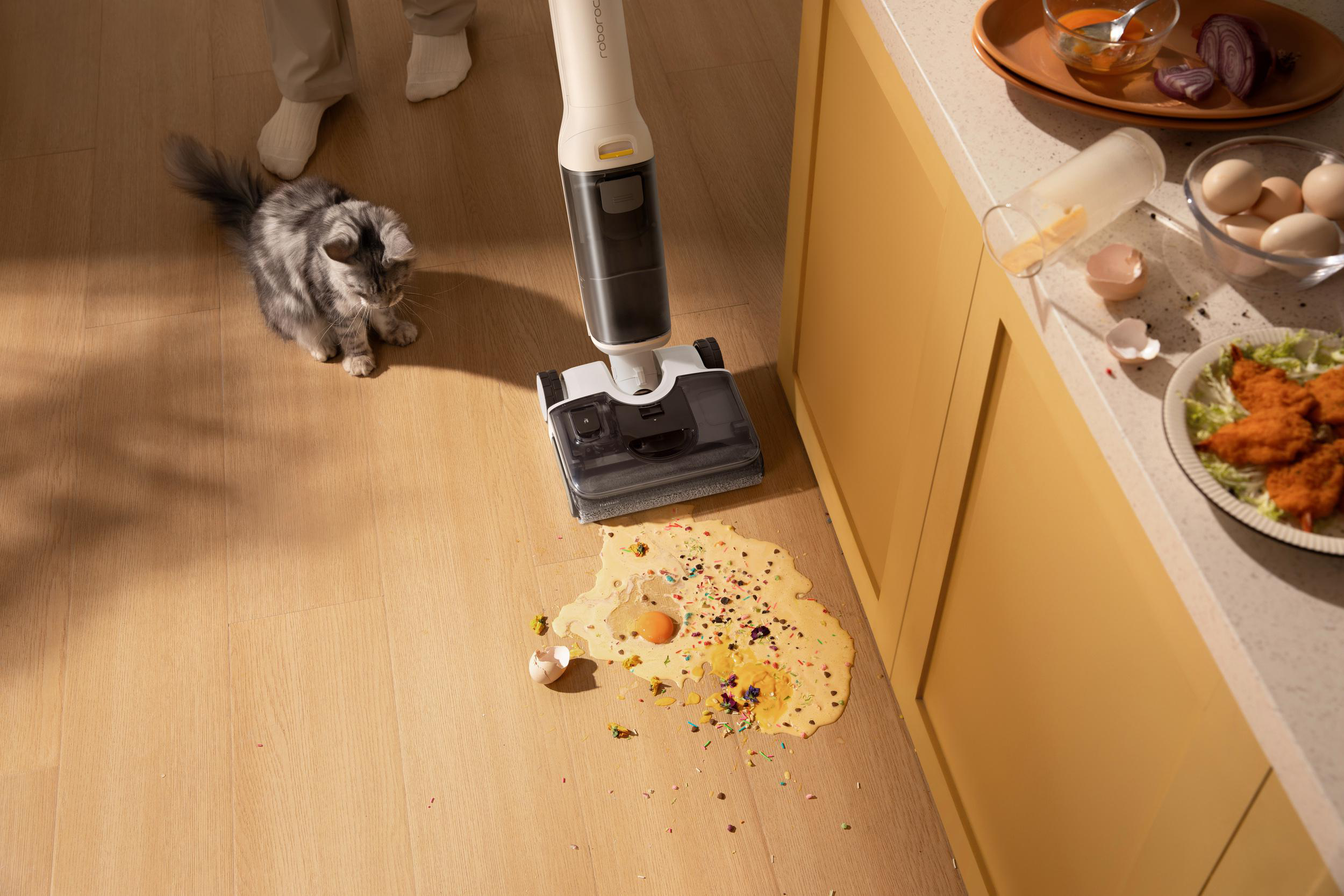 Eine Katze und ein Staubsauger auf einem Holzboden mit verschütteten Eiern und Streuseln.