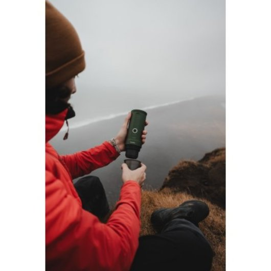 Persona con chaqueta roja filtra agua con dispositivo verde al aire libre en día de niebla.