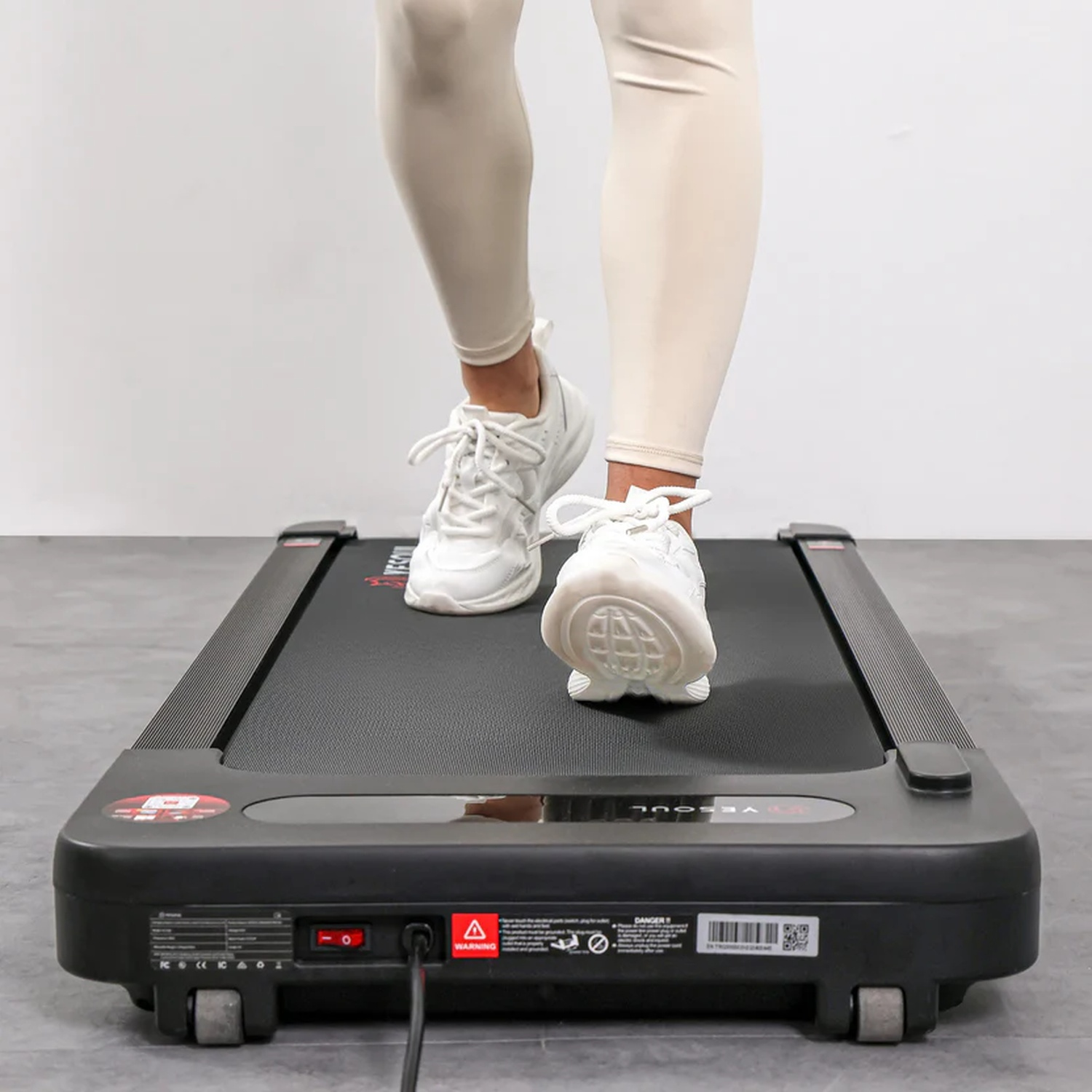 Una persona con leggings beige chiaro e scarpe da ginnastica bianche cammina su un tapis roulant nero.