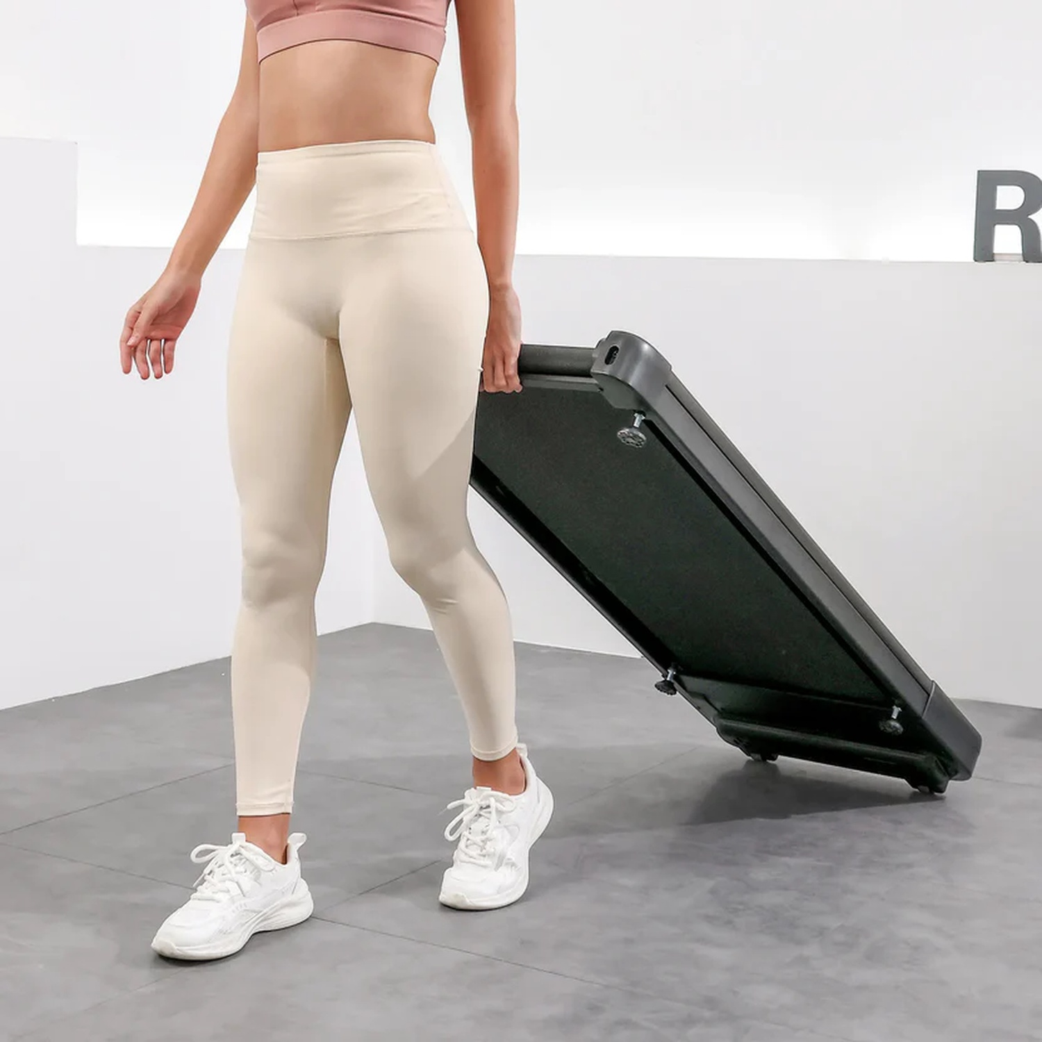 Una donna in abbigliamento sportivo beige porta un tapis roulant nero.