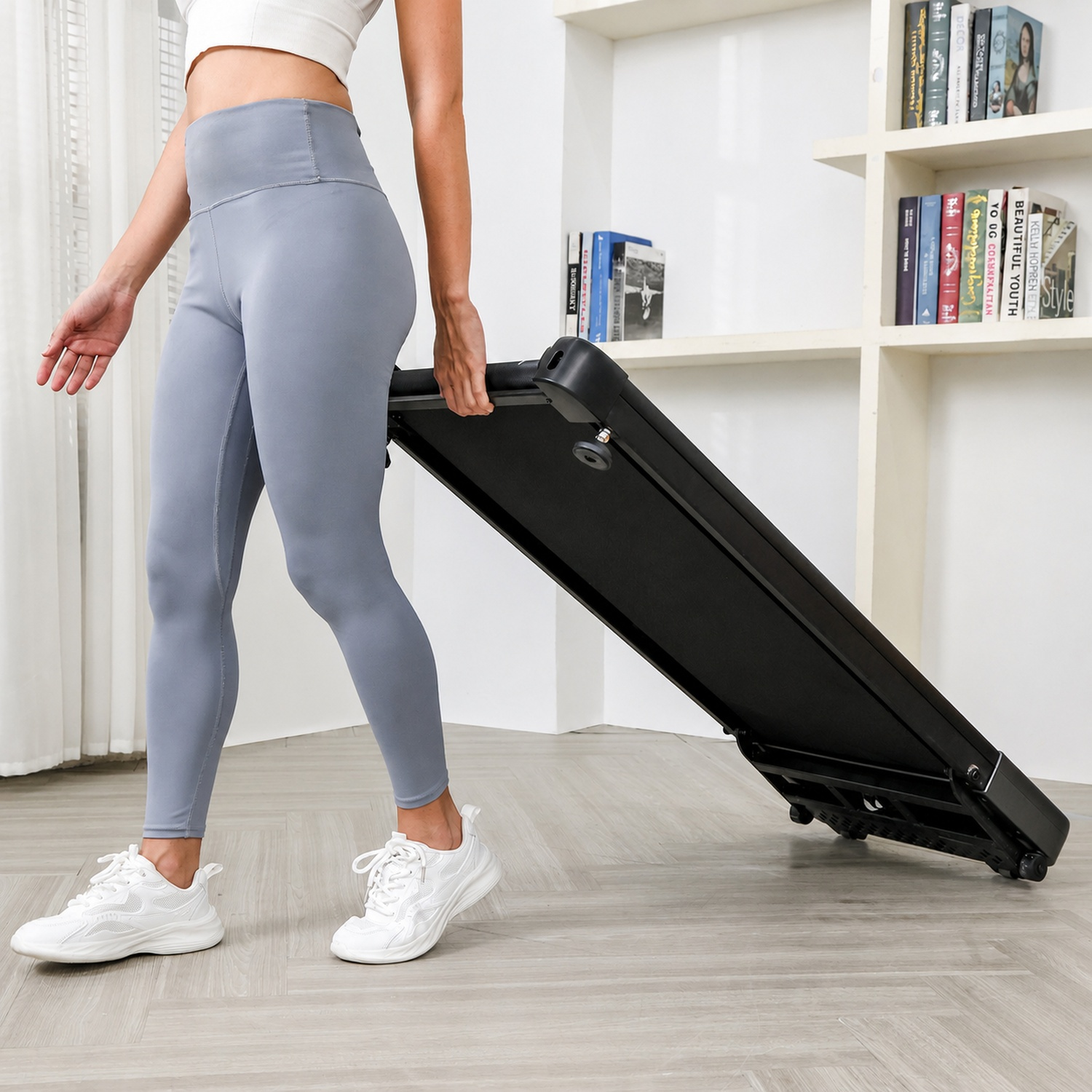 Una donna in leggings grigi e scarpe da ginnastica bianche porta un tapis roulant nero.
