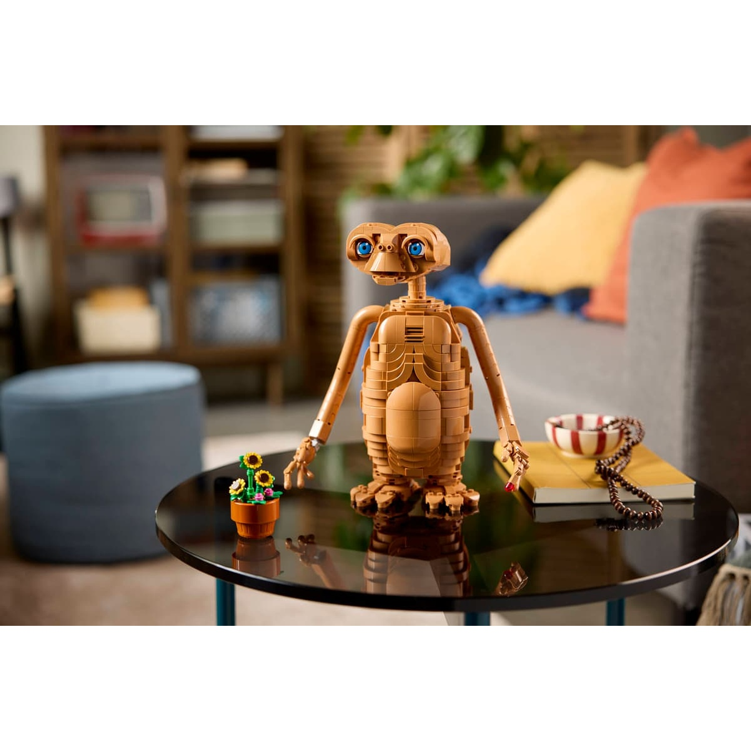Una figura marrone LEGO di E.T. si trova su un tavolo rotondo vicino a un vaso di girasoli.