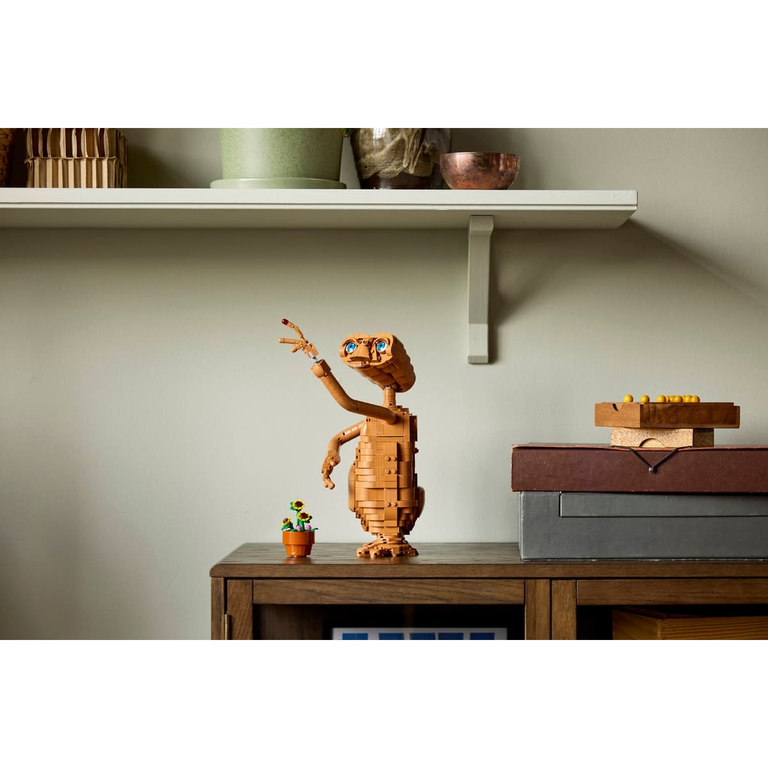Un ET Lego arancione con occhi blu sta accanto a una pianta in vaso su un mobile di legno.