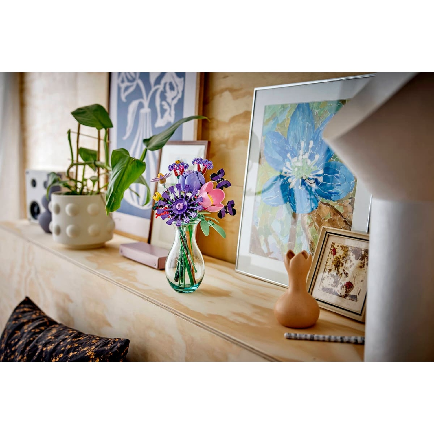 Un vaso di vetro con fiori viola e rosa si trova su una mensola di legno con piante e arte.
