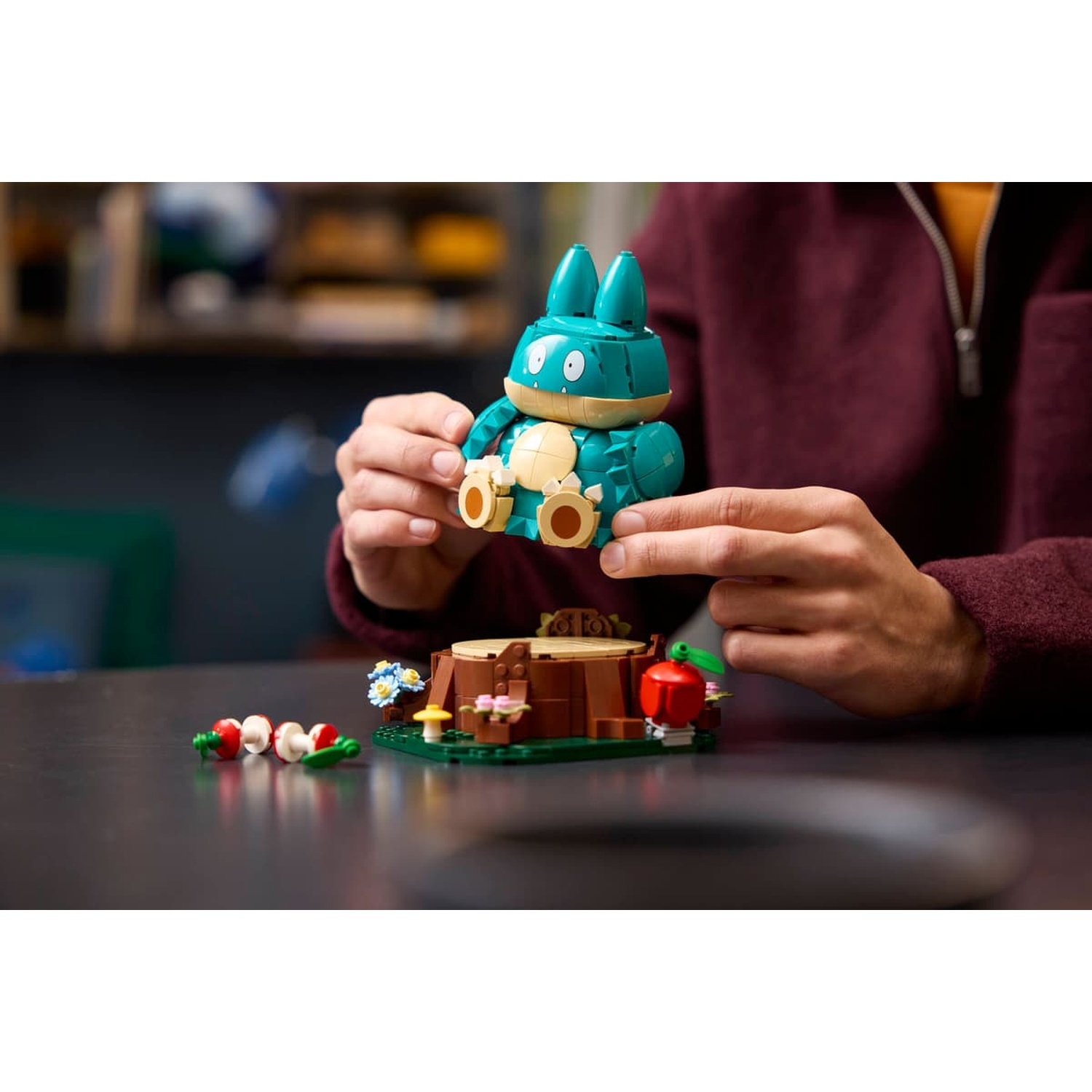 Una persona tiene un Pokémon Lego verde acqua su una base a tema forestale con funghi e una mela.