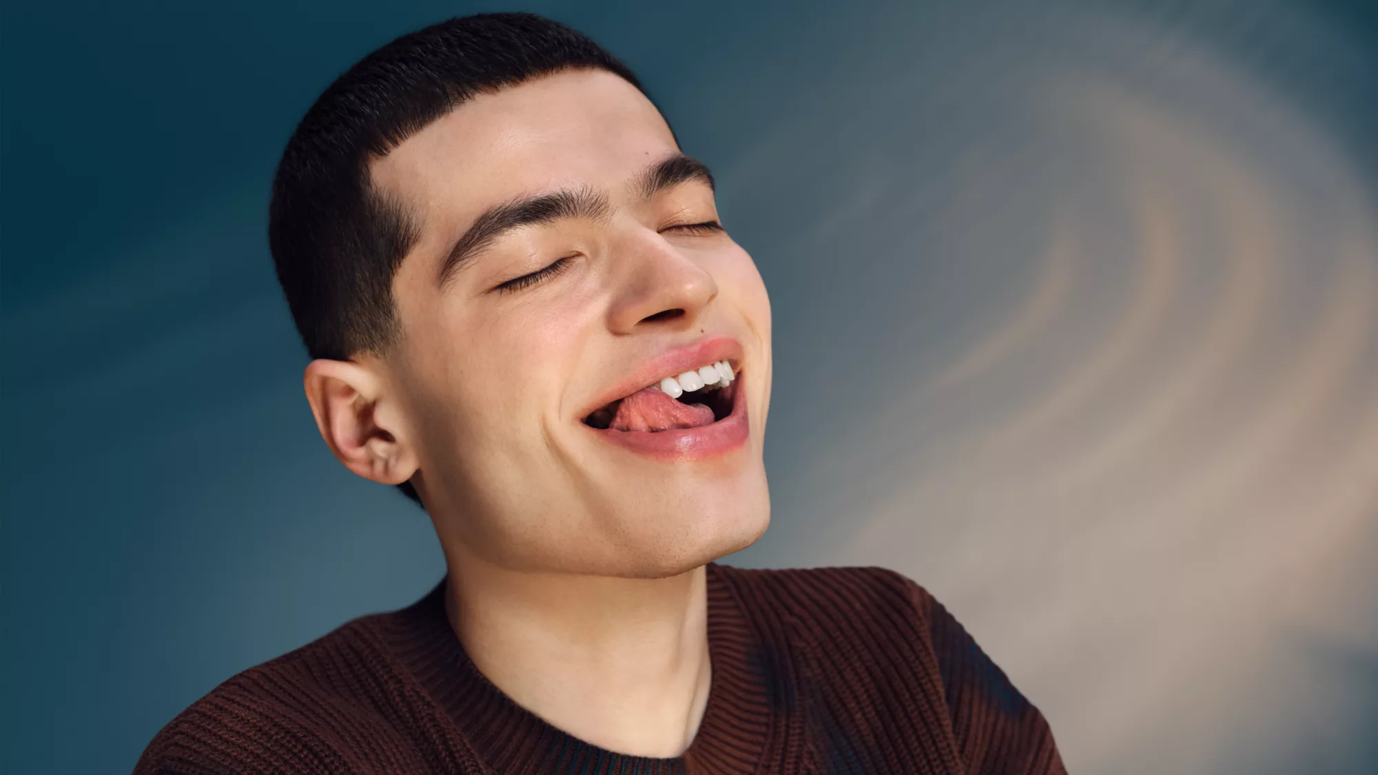 Ein Mann mit geschlossenen Augen und herausgestreckter Zunge, trägt einen braunen Pullover.