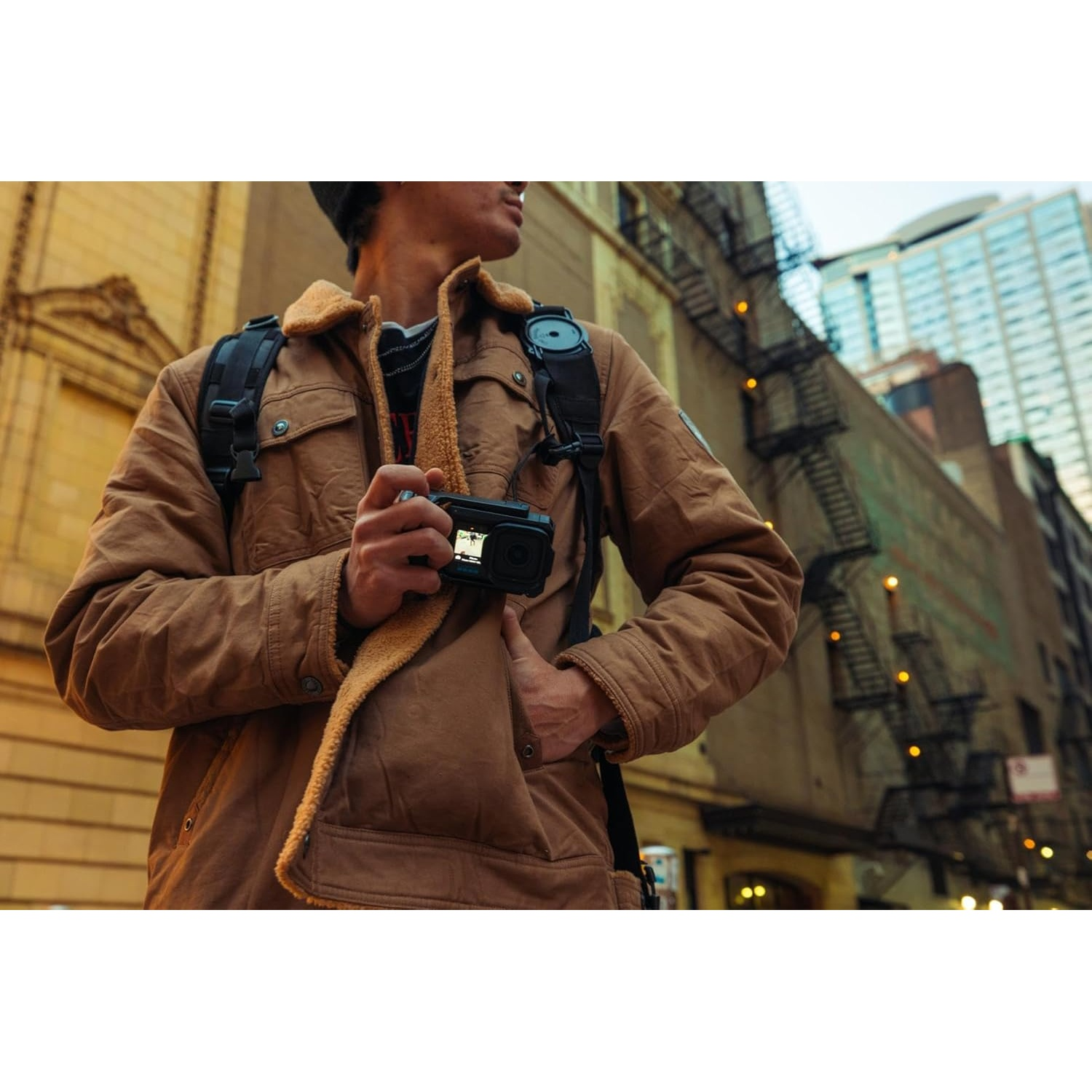 Man in brown jacket holding black camera on city street with buildings.