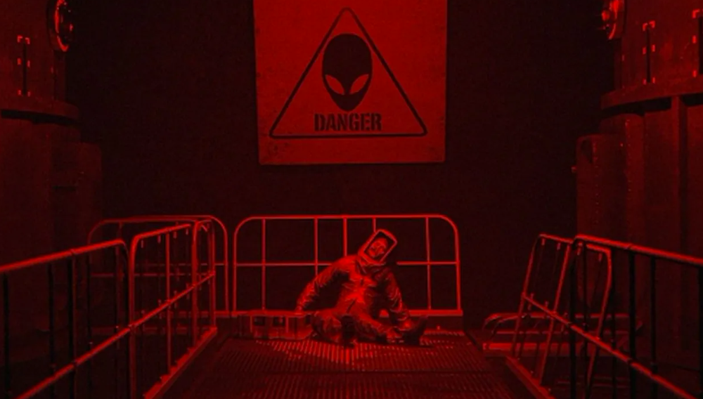 Ein Mann in einem Schutzanzug sitzt unter einem Warnschild mit einem Alien-Kopf.
