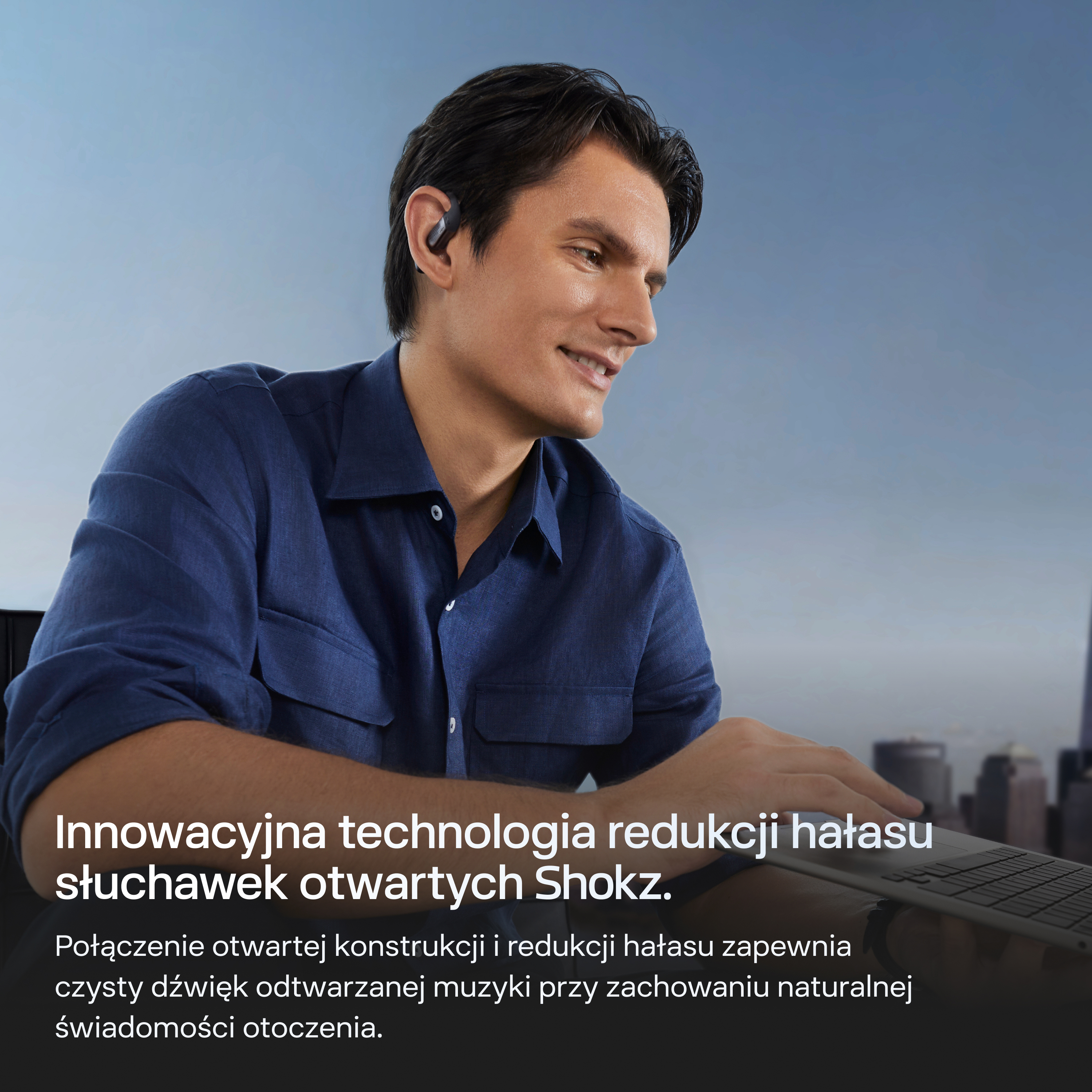 Mężczyzna ze słuchawkami używa laptopa, w tle panorama miasta.