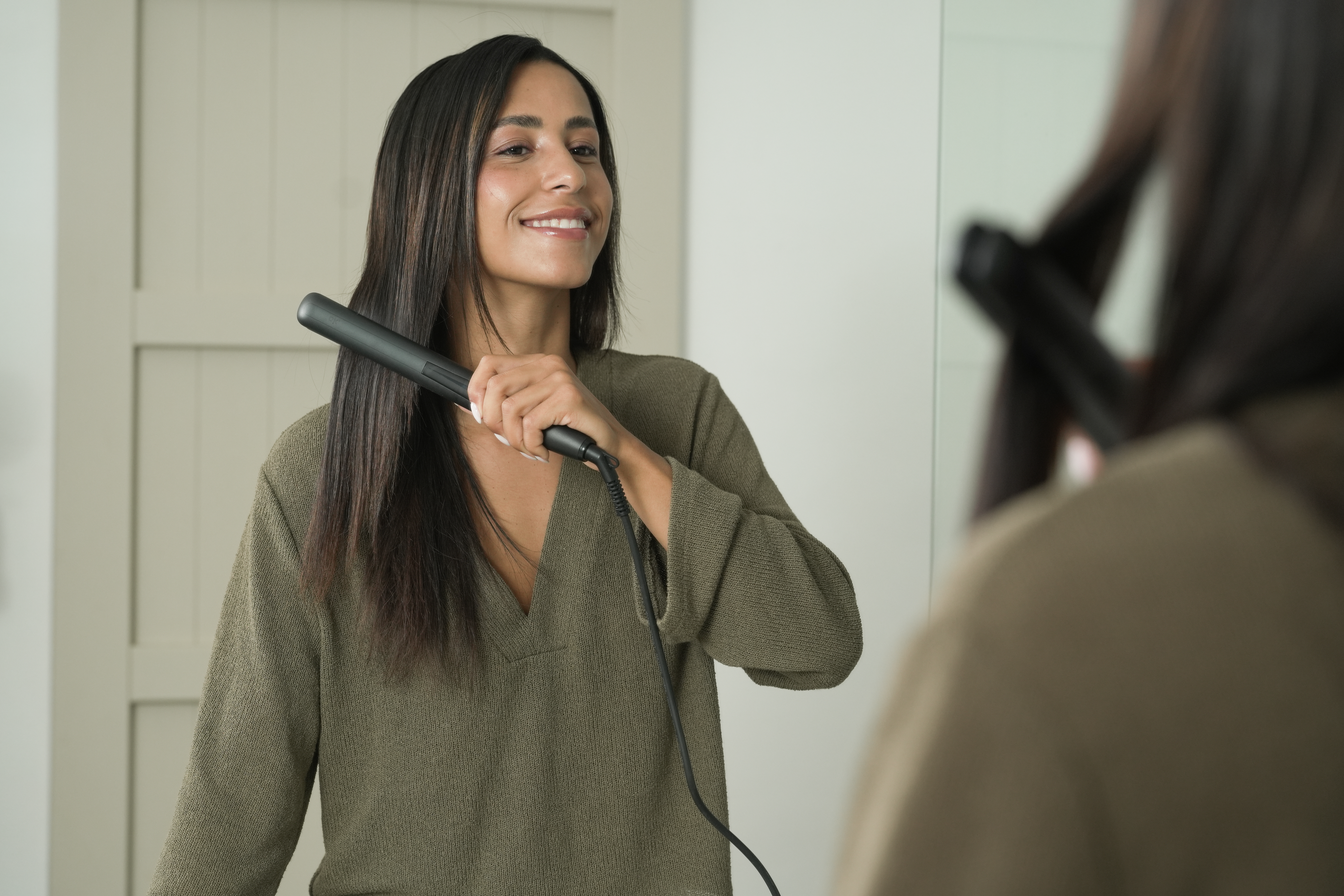Una mujer se alisa el pelo oscuro con una plancha negra, mirándose en un espejo.