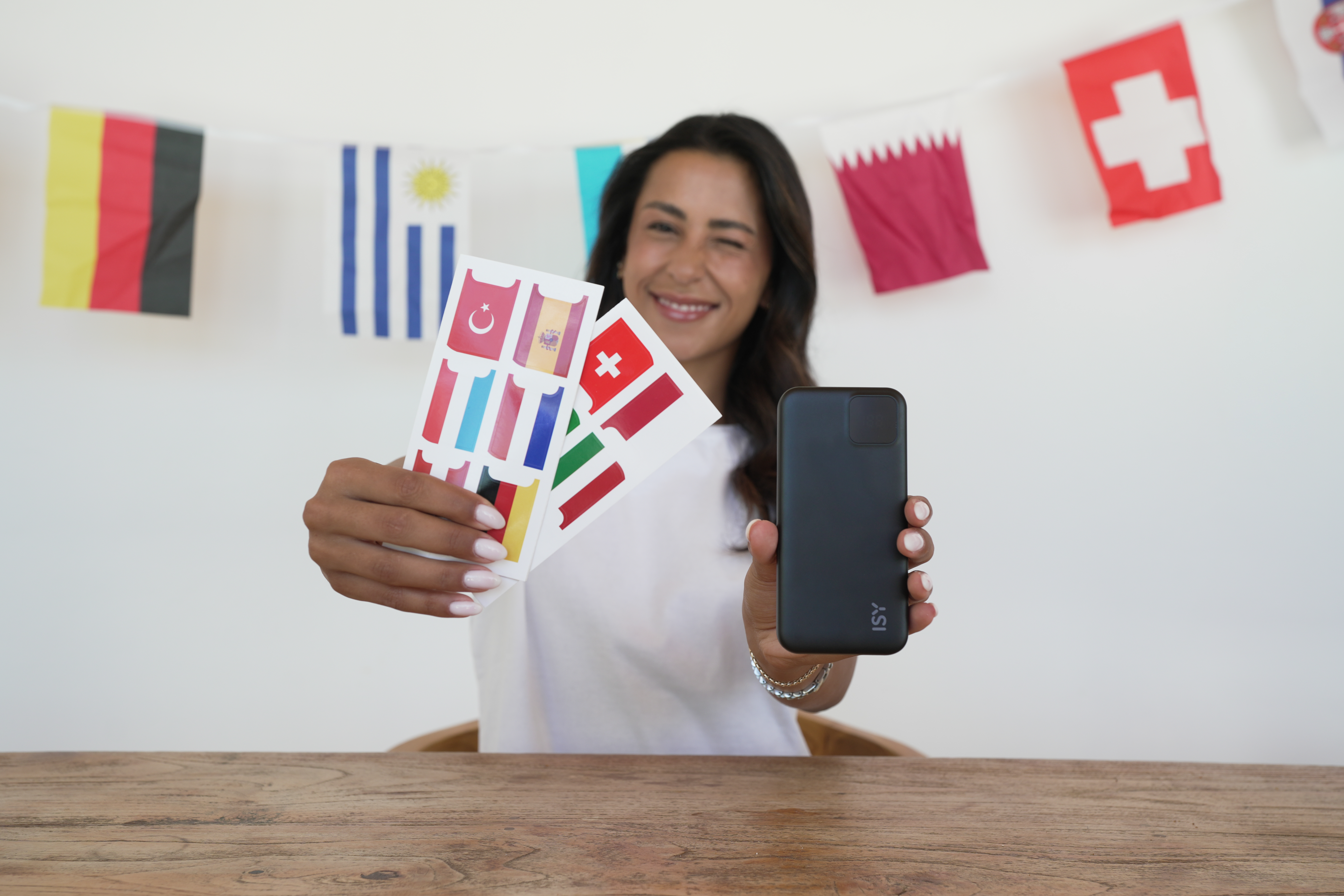 Frau hält Handy und Flaggen-Aufkleber vor Wimpelketten mit Länderflaggen.