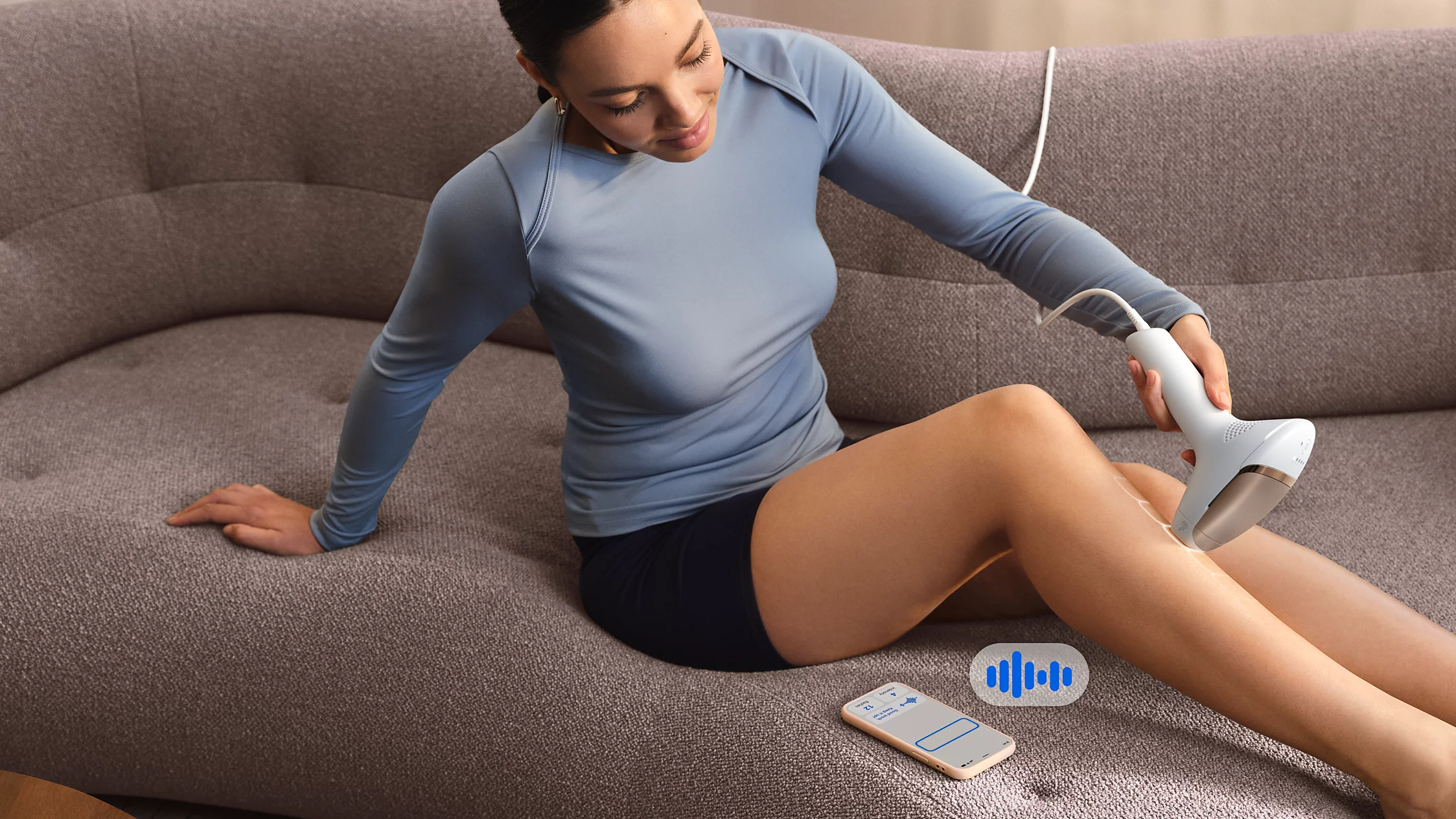 Frau auf Sofa behandelt Bein mit IPL-Gerät, Handy daneben.